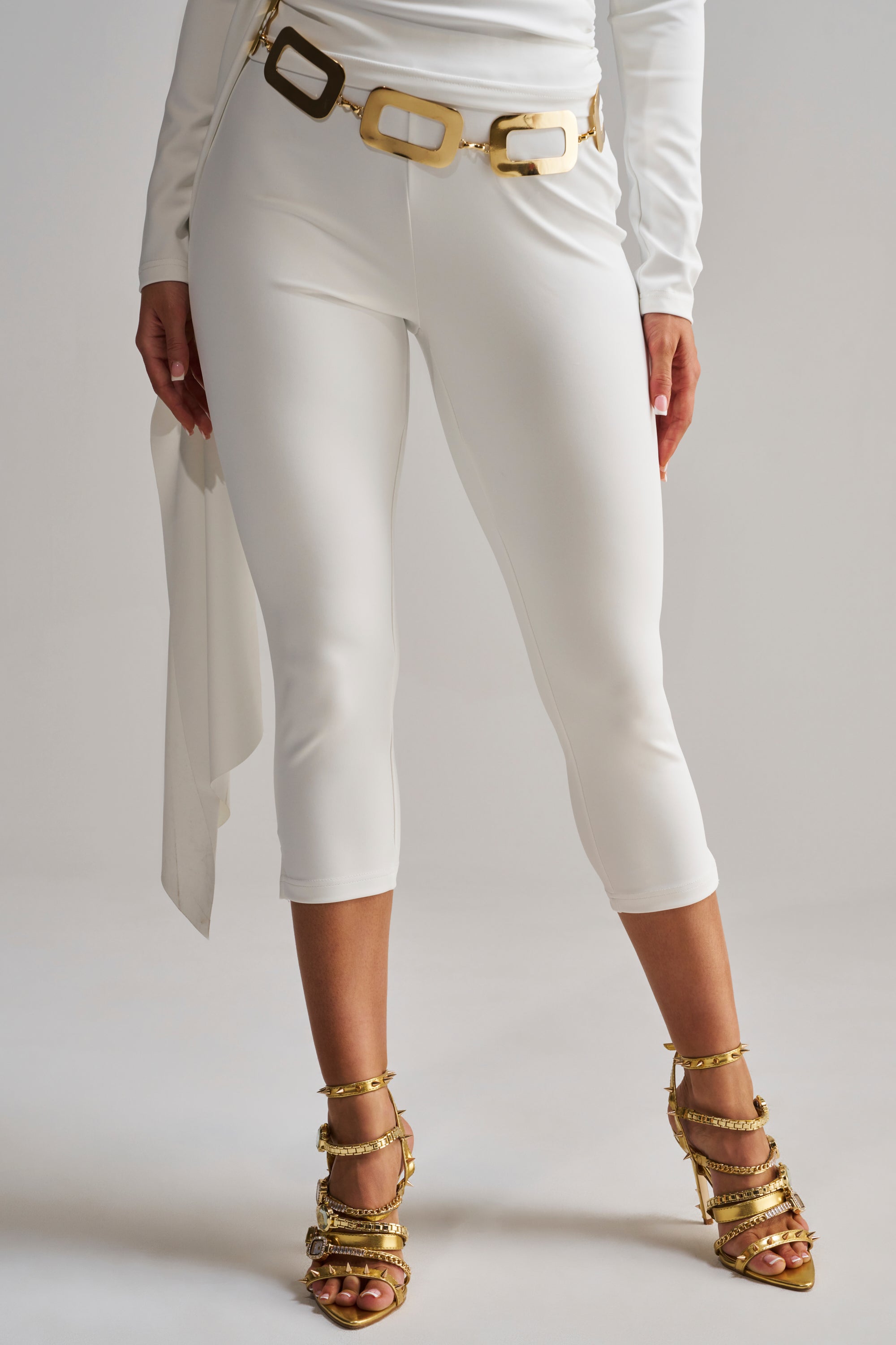 Wearing the MODEL OFF DUTY CAPRI in white, paired with a fitted long-sleeve top, a gold and white statement belt, and gold embellished high-heeled sandals for a modern, stylish look.