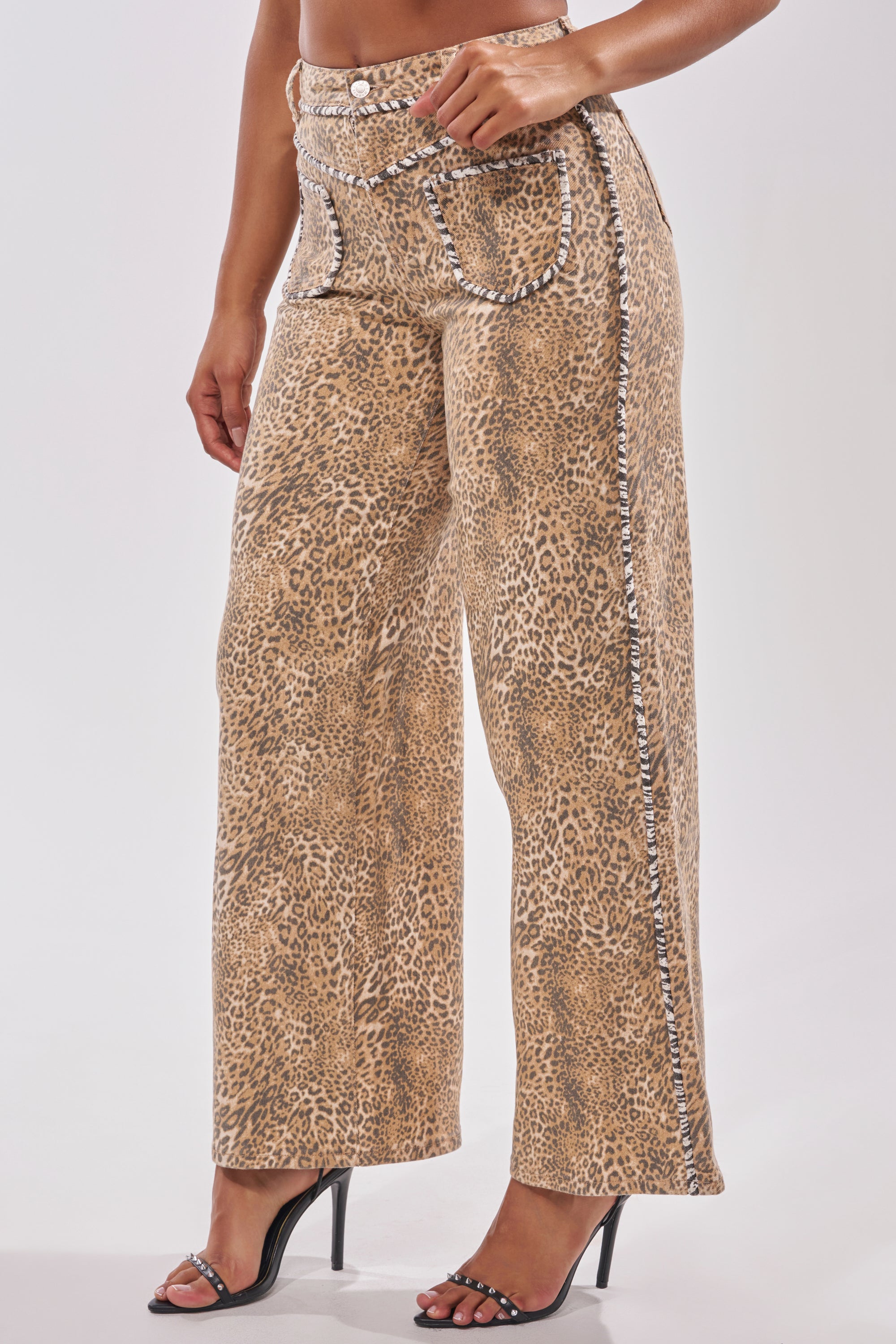 A person models IN THE WILD LEOPARD DENIM, featuring a high-waisted, wide leg silhouette with rhinestone trim, styled with black high-heeled sandals. Photo is cropped above the shoulders on a plain white background. 2