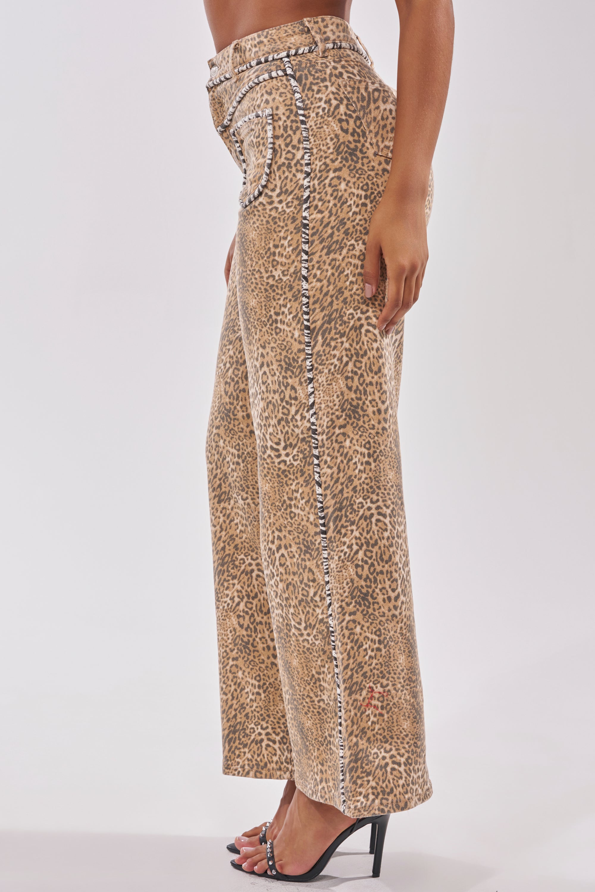 A person seen from the side wears IN THE WILD LEOPARD DENIM—high-waisted, wide leg leopard print jeans with zebra-print seam trim—and black open-toe high-heeled sandals. 3