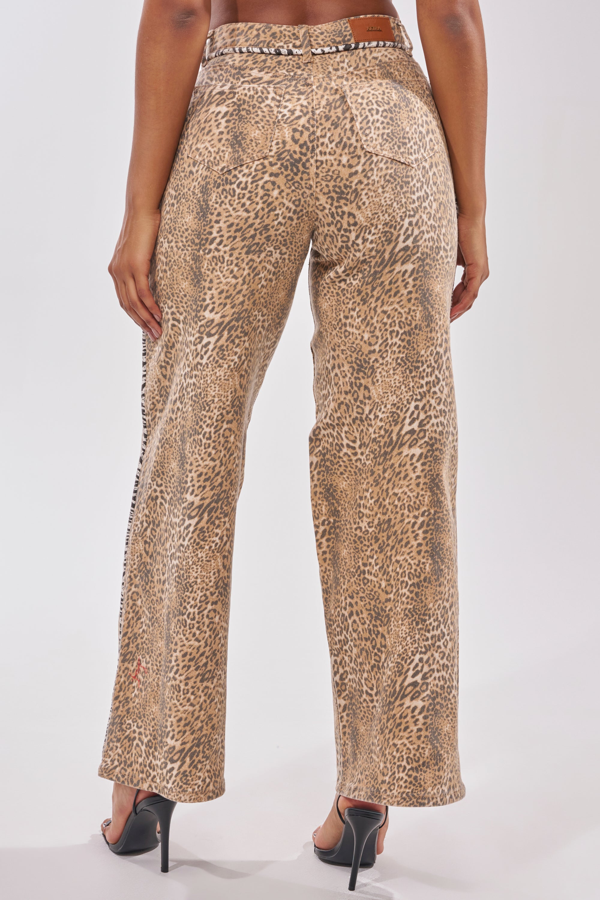 Someone shown from the back wears the IN THE WILD LEOPARD DENIM, a high-waisted, wide-leg leopard print pant, paired with high-heeled sandals against a plain white background. 4