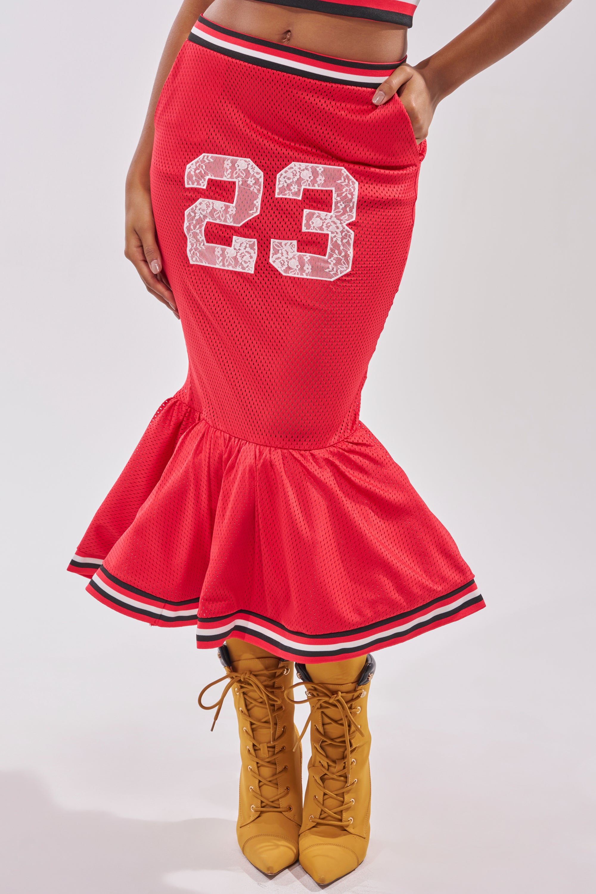 A person sports the bold GAME CHANGER PEPLUM MIDI SKIRT in red with a white 23 and black-and-white striped hem, styled with tan lace-up boots for a standout courtside streetwear vibe. 1