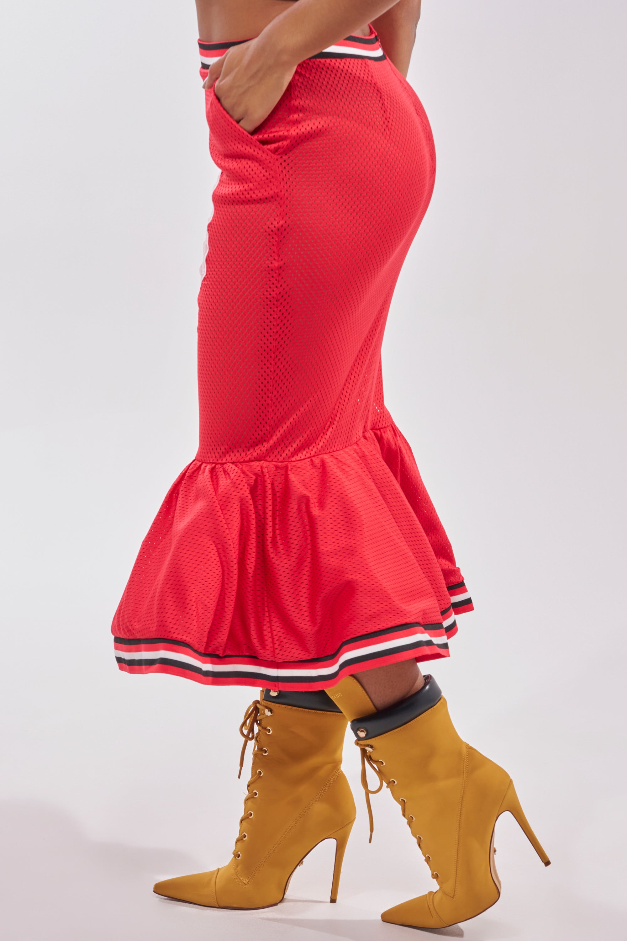 Someone wears the GAME CHANGER PEPLUM MIDI SKIRT, a fitted, flared red jersey piece with striped trim at the hem and waistband, styled with mustard yellow lace-up high-heeled boots for a bold courtside streetwear vibe. Upper body mostly out of frame. 2