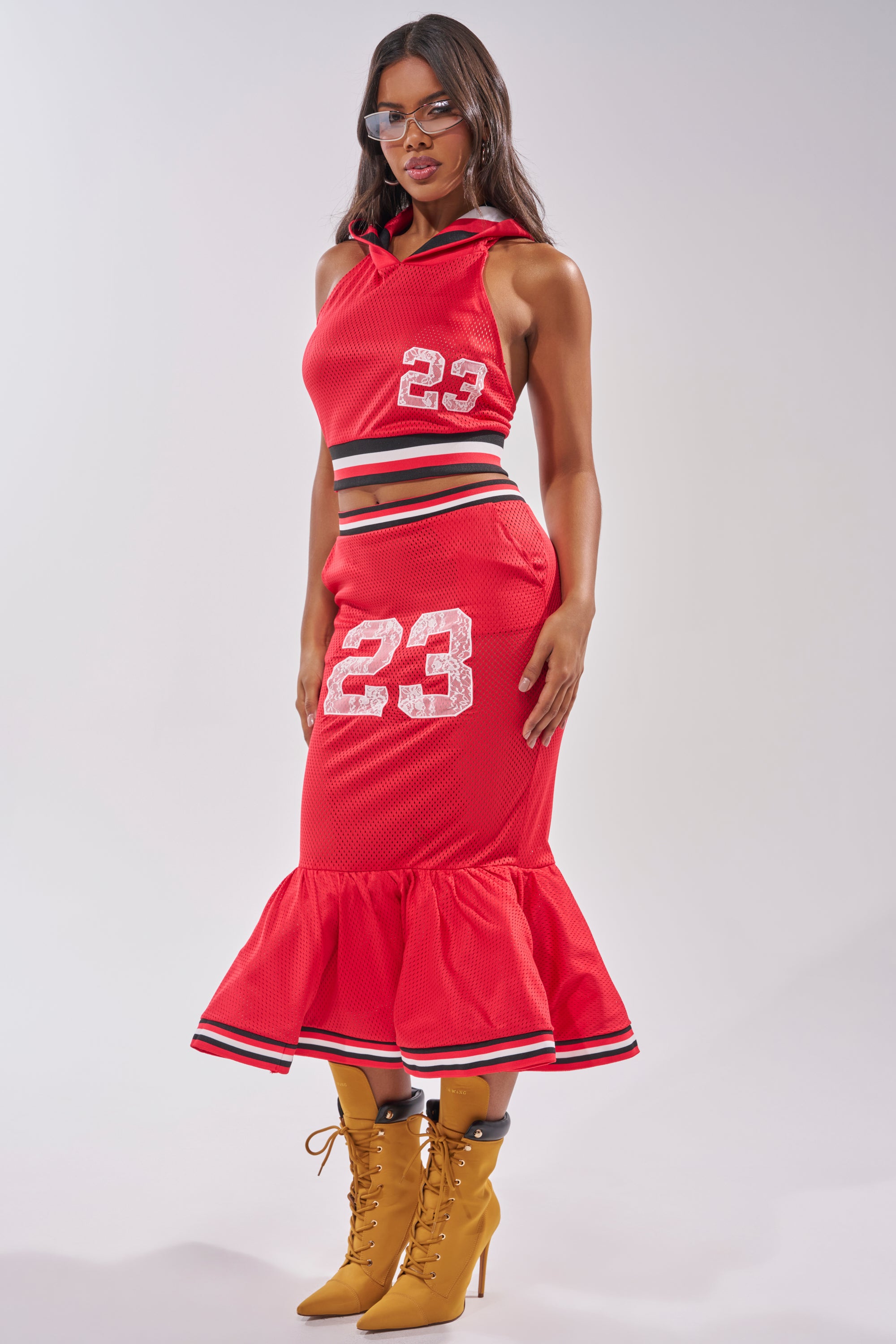 A woman serves courtside style in a sleeveless red crop hoodie and the GAME CHANGER PEPLUM MIDI SKIRT featuring the number "23," paired with sunglasses and tan lace-up heeled boots. 5