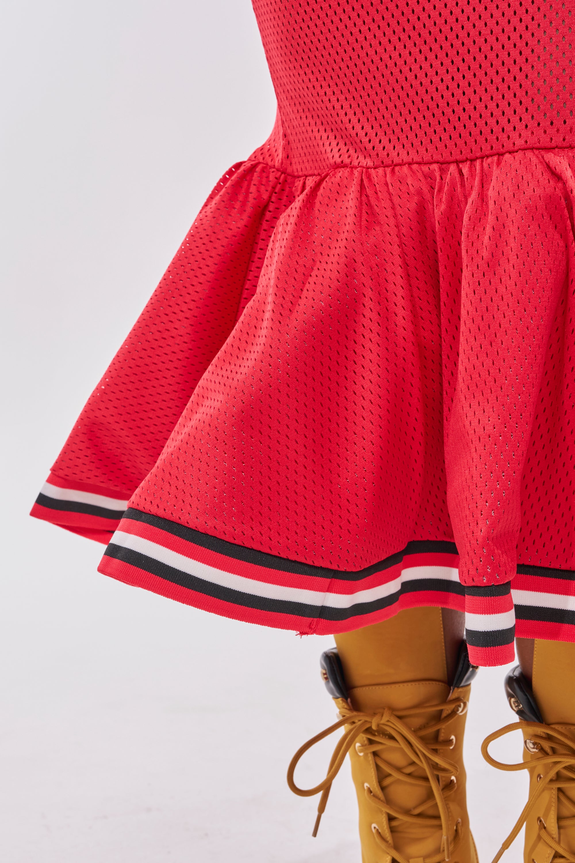 Wearing the GAME CHANGER PEPLUM MIDI SKIRT with black, white, and red striped trim and yellow lace-up boots, the photo is cropped to highlight just the skirt’s lower half and shoes for a courtside streetwear look. 8