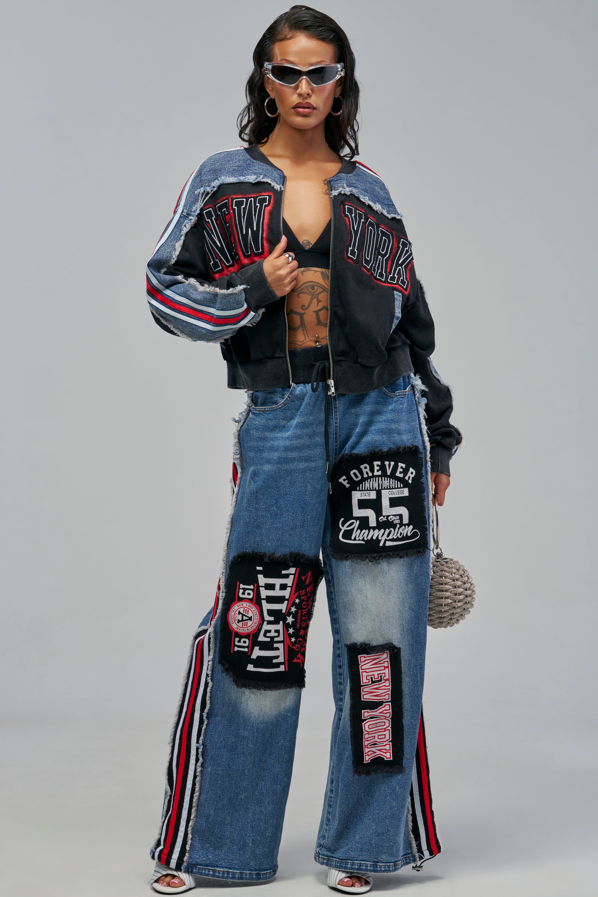 A model wearing a CHAMPION PATCHWORK DENIM cropped bomber jacket with “NEW YORK” lettering and matching wide-leg graphic jeans featuring sports-themed patches and side stripes.