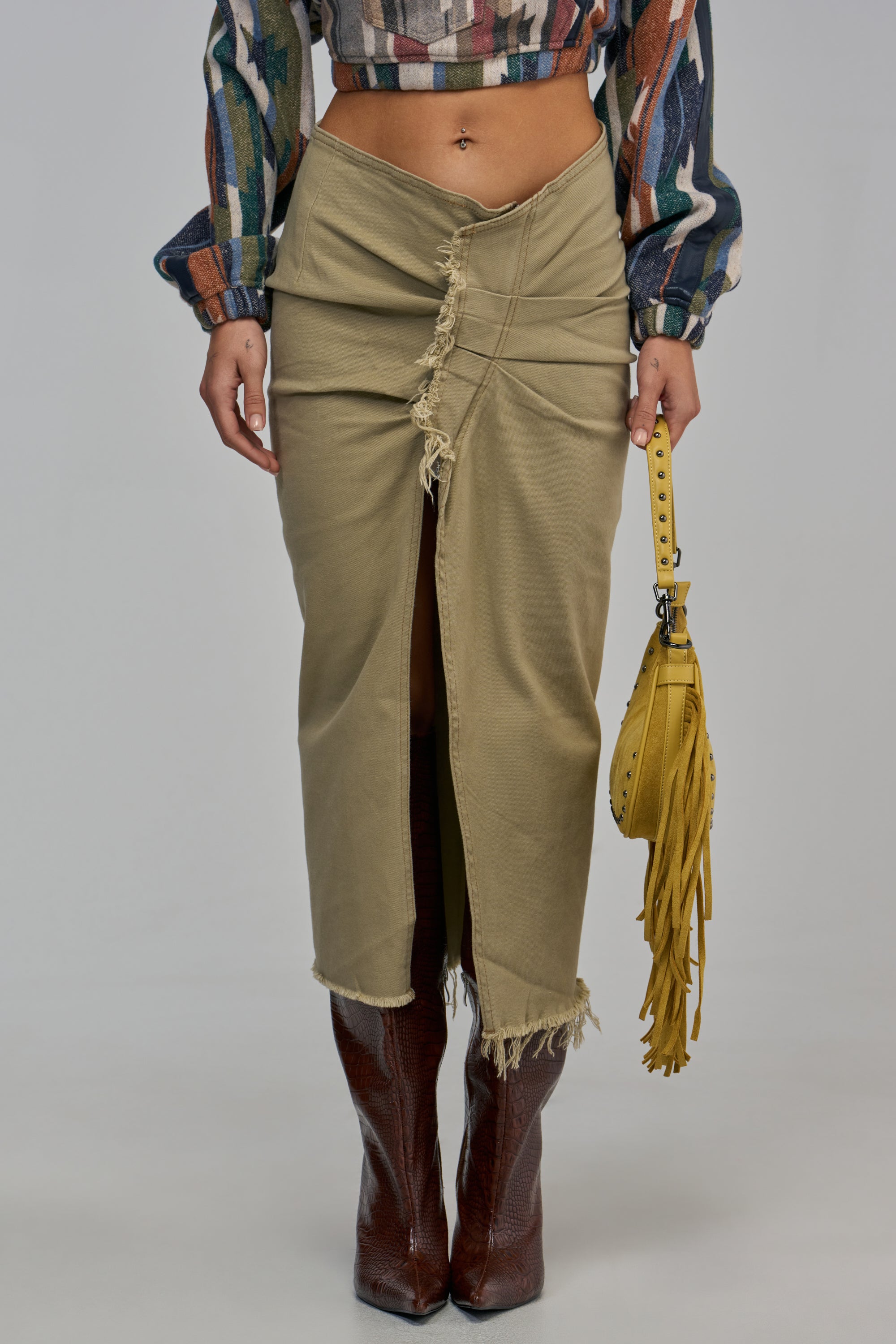 IM YOUR FAVORITE DENIM SKIRT in olive green khaki with a front slit, raw hem, and exposed stitching, worn with a patterned cropped long-sleeve top, brown knee-high boots, and a yellow fringed handbag.