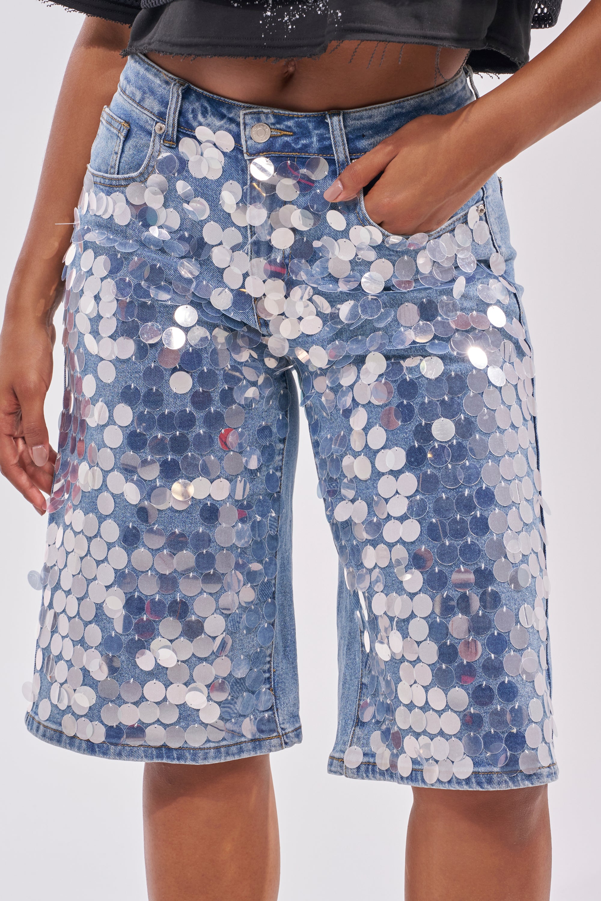 Wearing the SPARKLE AND SHINE BERMUDA SHORT, covered in large, shiny silver sequins, someone stands with one hand in their pocket—a bold choice for standout festival style. 1