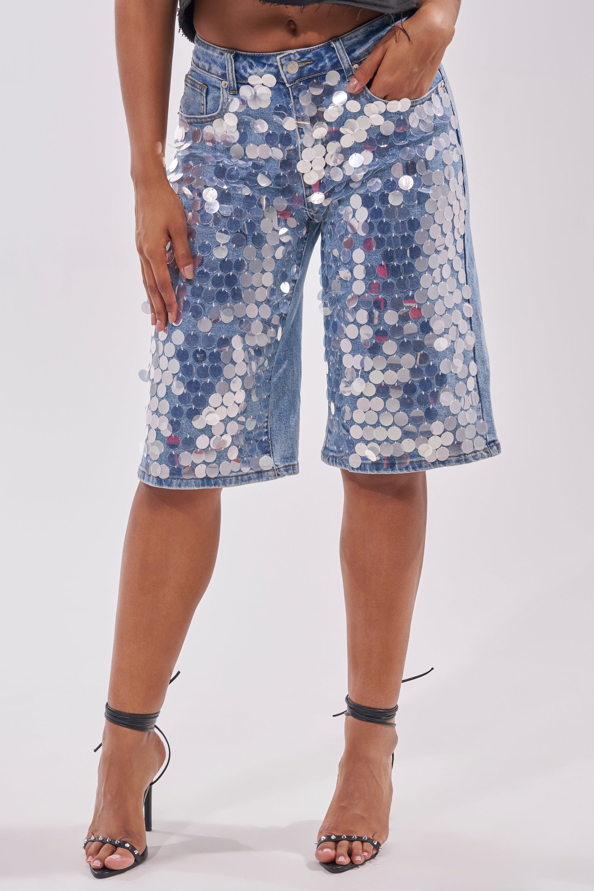 Wearing the SPARKLE AND SHINE BERMUDA SHORT, adorned with large silver sequins, and black strappy heels, a person poses with a hand in their pocket for a festival-ready look against a plain white background. 2