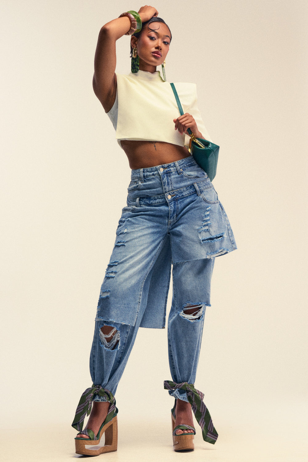 Wearing the TOO COOL FOR YOU RELAXED DENIM, this woman showcases an effortlessly chic streetwear look, pairing the wide-leg distressed jeans—featuring layered denim and scarf details at the ankles—with a white crop top, platform sandals, green earrings, bangles, and a matching green shoulder bag as she poses confidently against a plain background.