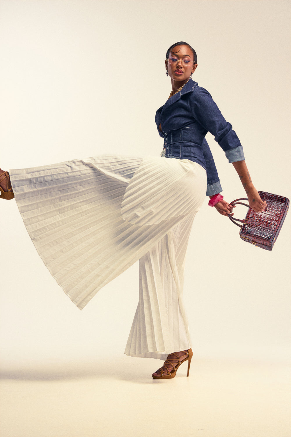 A woman poses stylishly in a denim jacket, DRIFTING AWAY PLEATED TROUSER high-rise wide-leg pants, brown heels, and glasses, holding a textured handbag with one leg playfully raised against a neutral background.