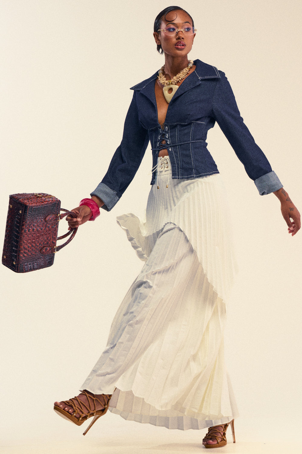 A person in a denim corset jacket, DRIFTING AWAY PLEATED TROUSER, brown strappy heels, statement necklace, pink bracelet, and glasses walks while holding a textured brown handbag.