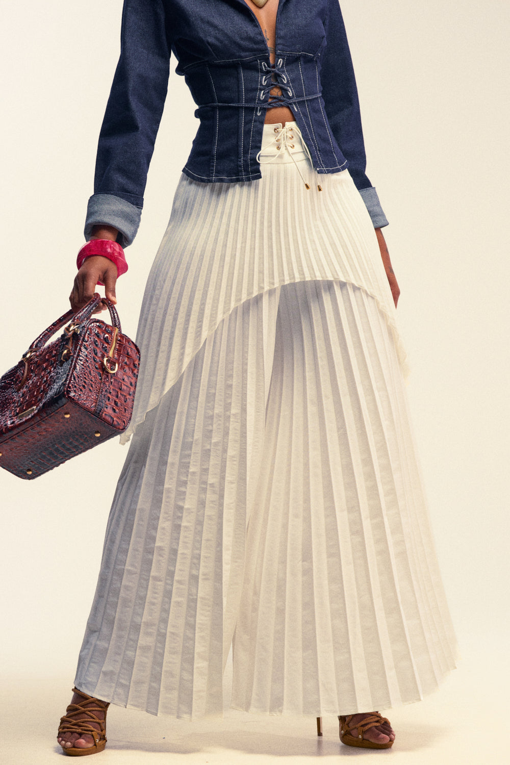 A person is wearing a denim lace-up top, DRIFTING AWAY PLEATED TROUSER in white, brown sandals, and holding a textured brown handbag.