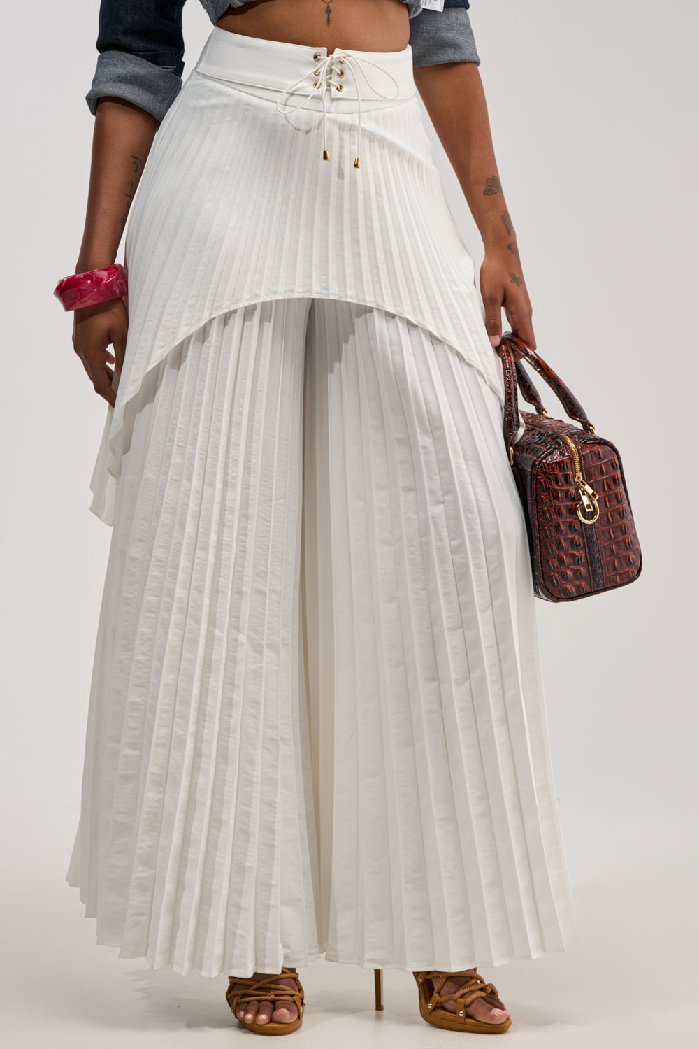 A woman wears the DRIFTING AWAY PLEATED TROUSER—high-rise, wide-leg pants with white pleated layers that resemble a skirt—paired with a cropped denim top, brown heeled sandals, a pink bracelet, and carries a brown textured handbag.