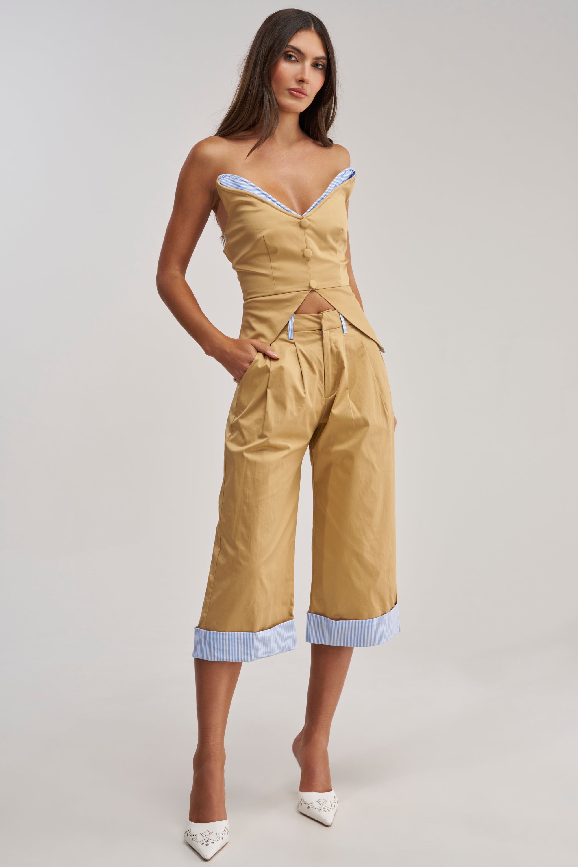 A woman stands confidently in the TOP TIER POPLIN TRIM PANT, tan culottes with light blue striped poplin trim, styled with an off-the-shoulder top and white pointed heels—an effortlessly chic spring look against a minimalist backdrop.