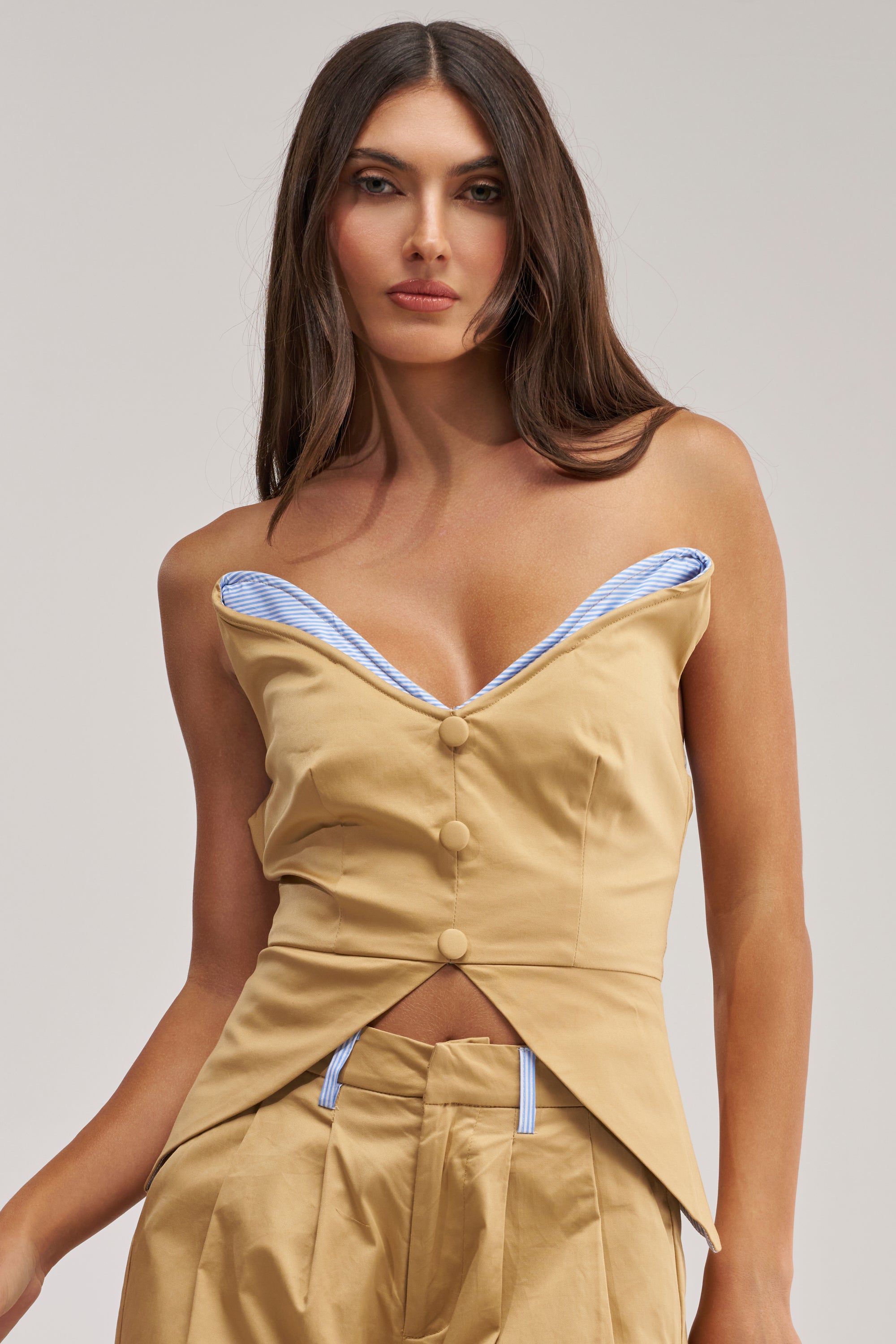 A woman with long brown hair and a neutral expression wears the TOP TIER POPLIN TRIM CORSET, a tan off-shoulder top with khaki corset details, blue striped poplin trim, folded neckline, angular hem, and matching high-waisted pants.