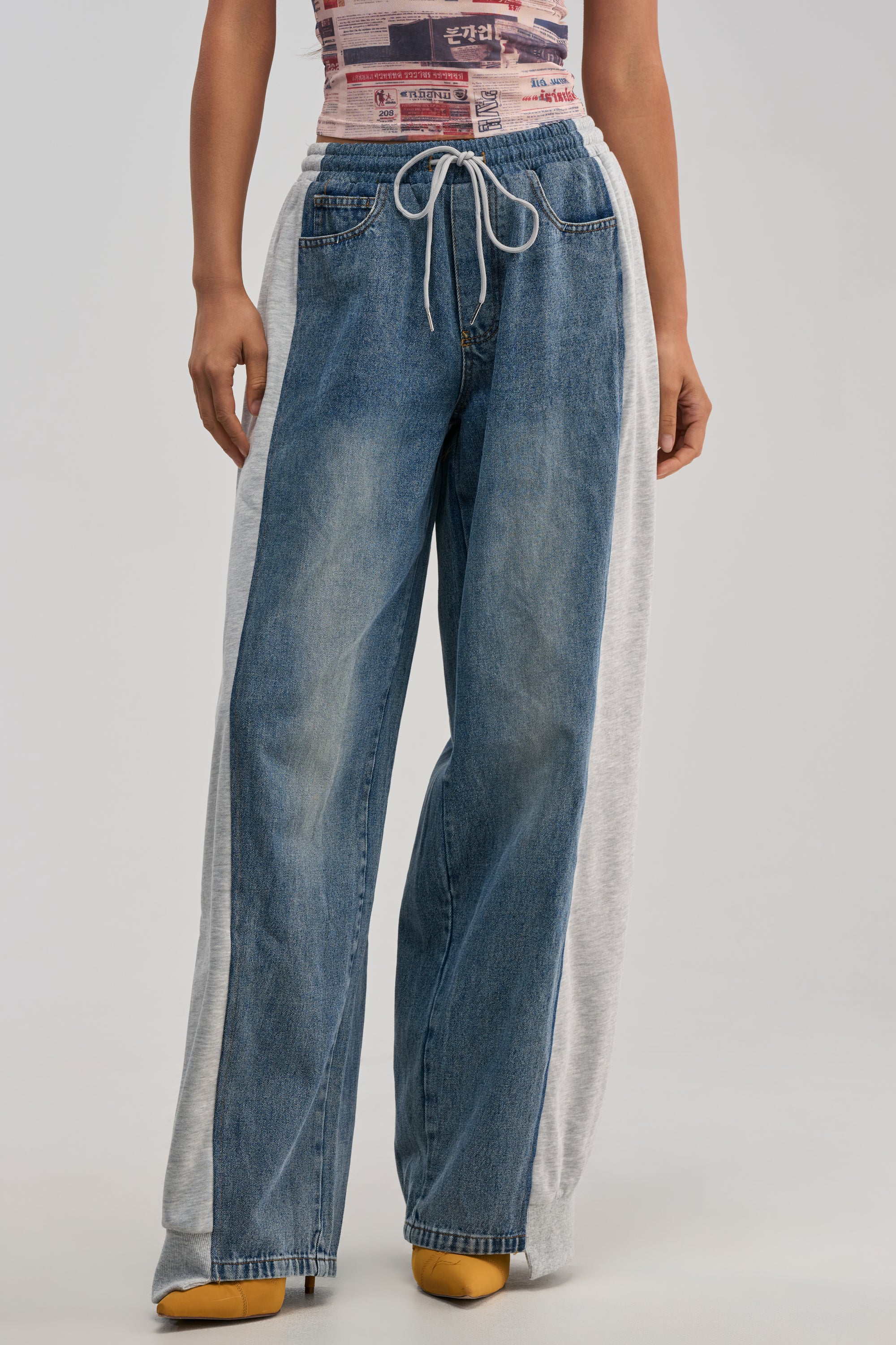 Wearing the COOL GIRL ERA JOGGER—wide-leg jeans featuring gray sweatpant panels on the sides and a drawstring waist for a denim jogger-inspired vibe—styled with a sleeveless printed top and yellow shoes. Captured from the waist down against a plain background, this look is the ultimate streetwear statement.