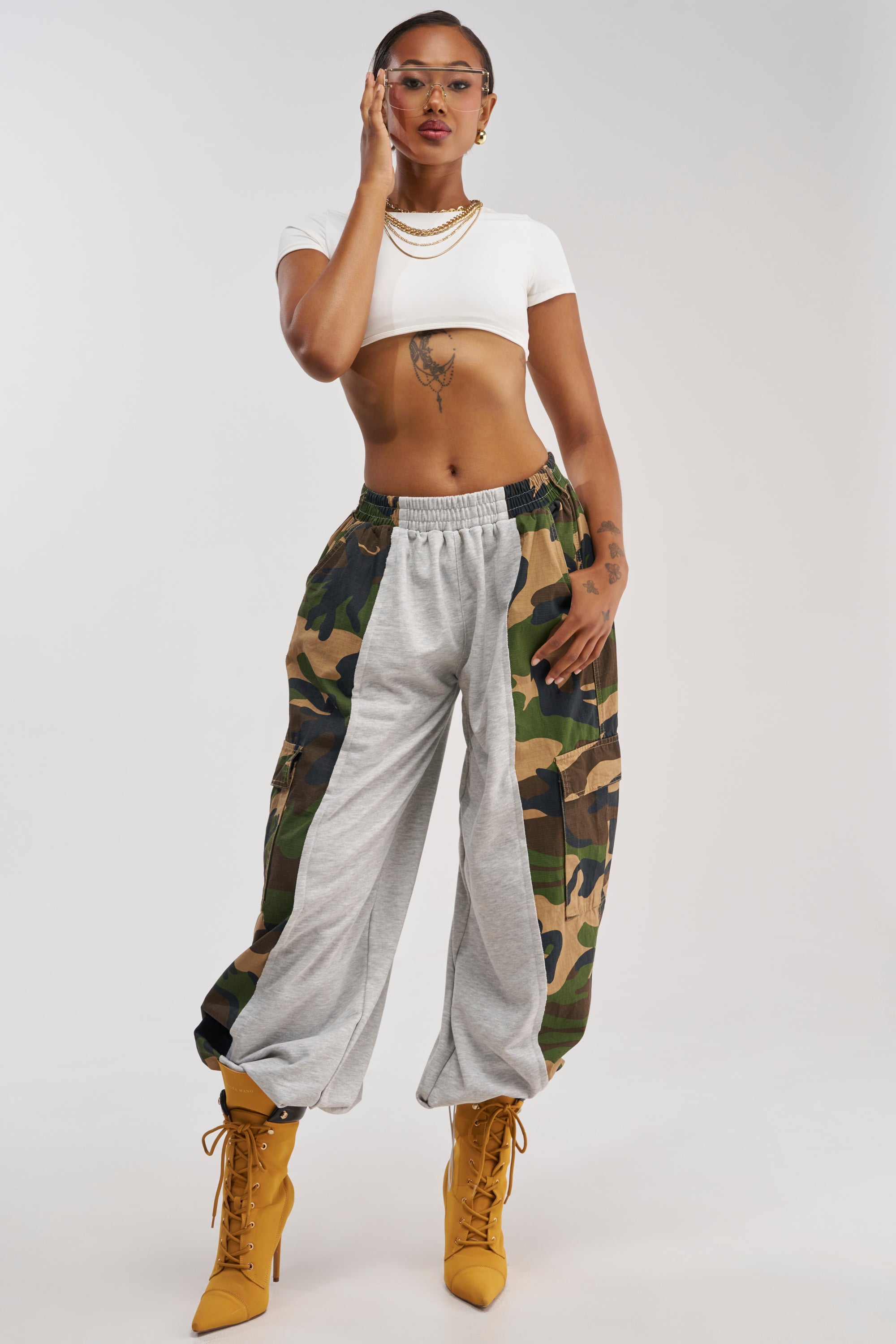 A woman poses confidently in the COOLEST ON THE BLOCK CAMO JOGGER—camo wide leg jogger pants with cargo pockets—paired with a white crop top and yellow heeled boots. She accessorizes with glasses, gold necklaces, and hoop earrings, her hand poised thoughtfully on her face, effortlessly demonstrating how loungewear can be styled chicly.