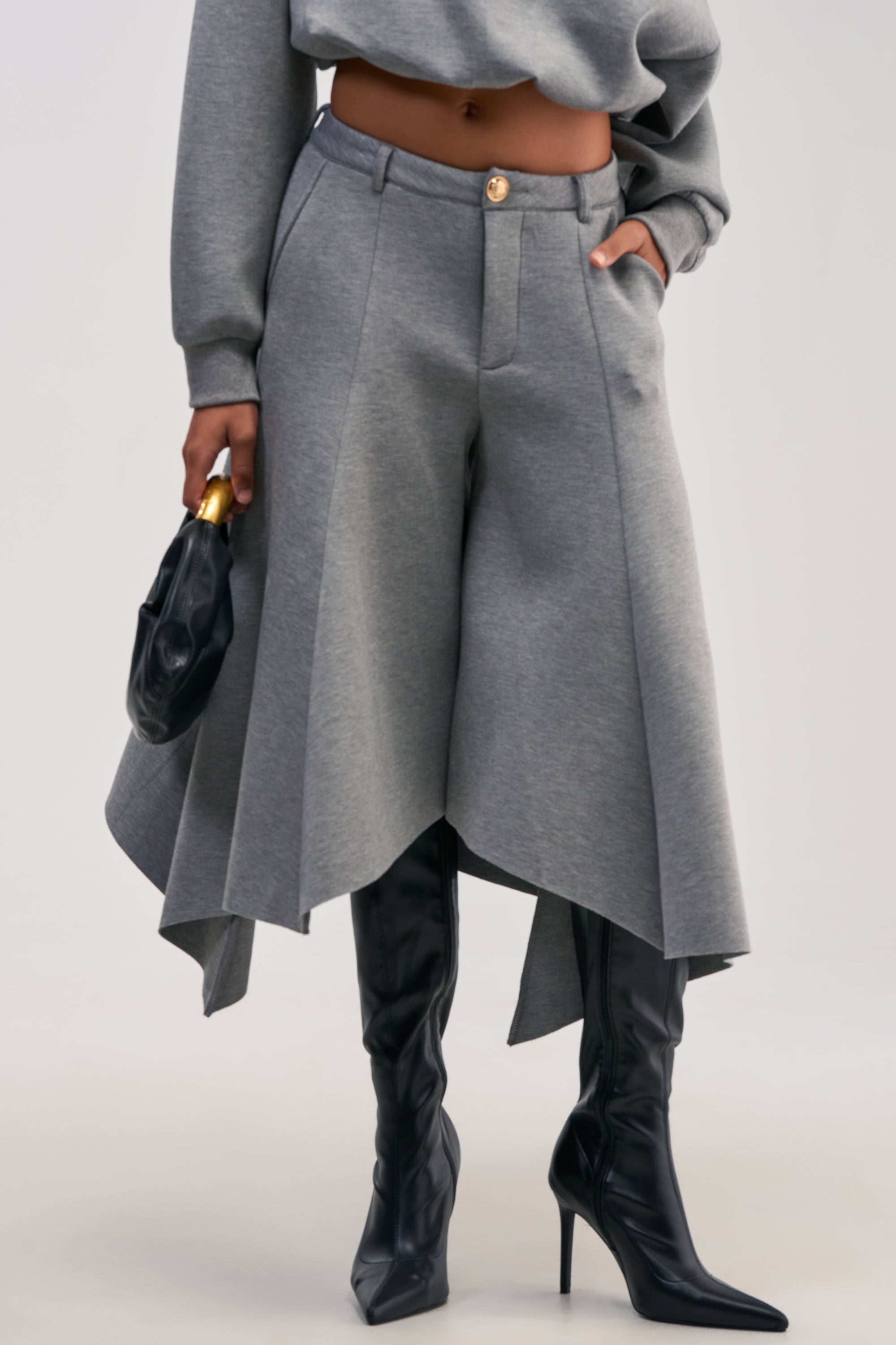 A person wearing the WILD AND FREE CULOTTES in grey, featuring a draped silhouette, cropped legs, and a high-rise waistband, styled with a matching gray top, black high-heeled boots, and holding a black clutch bag with a gold accent.