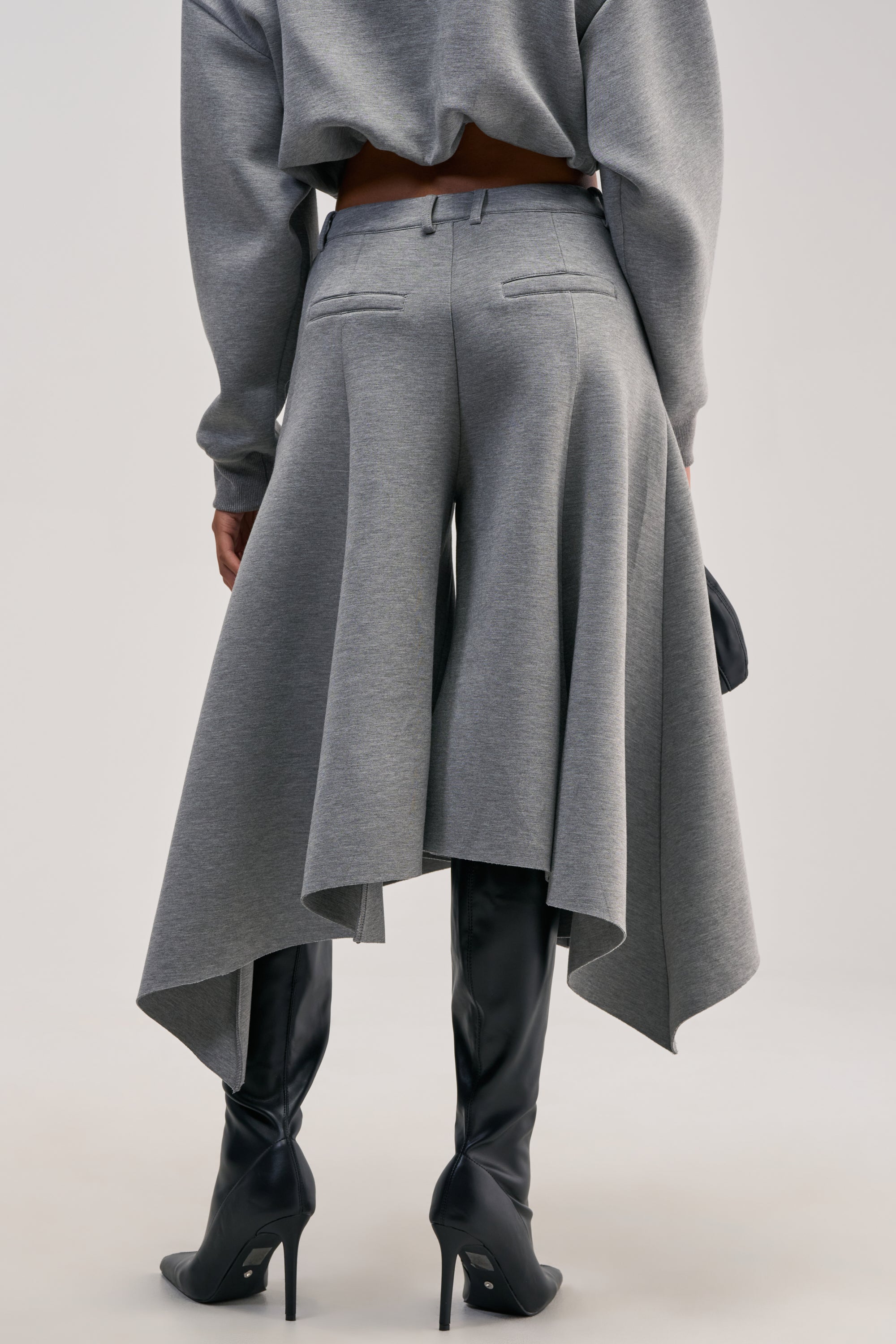 A person is shown from the back against a plain background wearing the WILD AND FREE CULOTTES—gray, asymmetrical skirt-pants featuring pointed hems and a high-rise waistband—paired with black high-heeled boots.