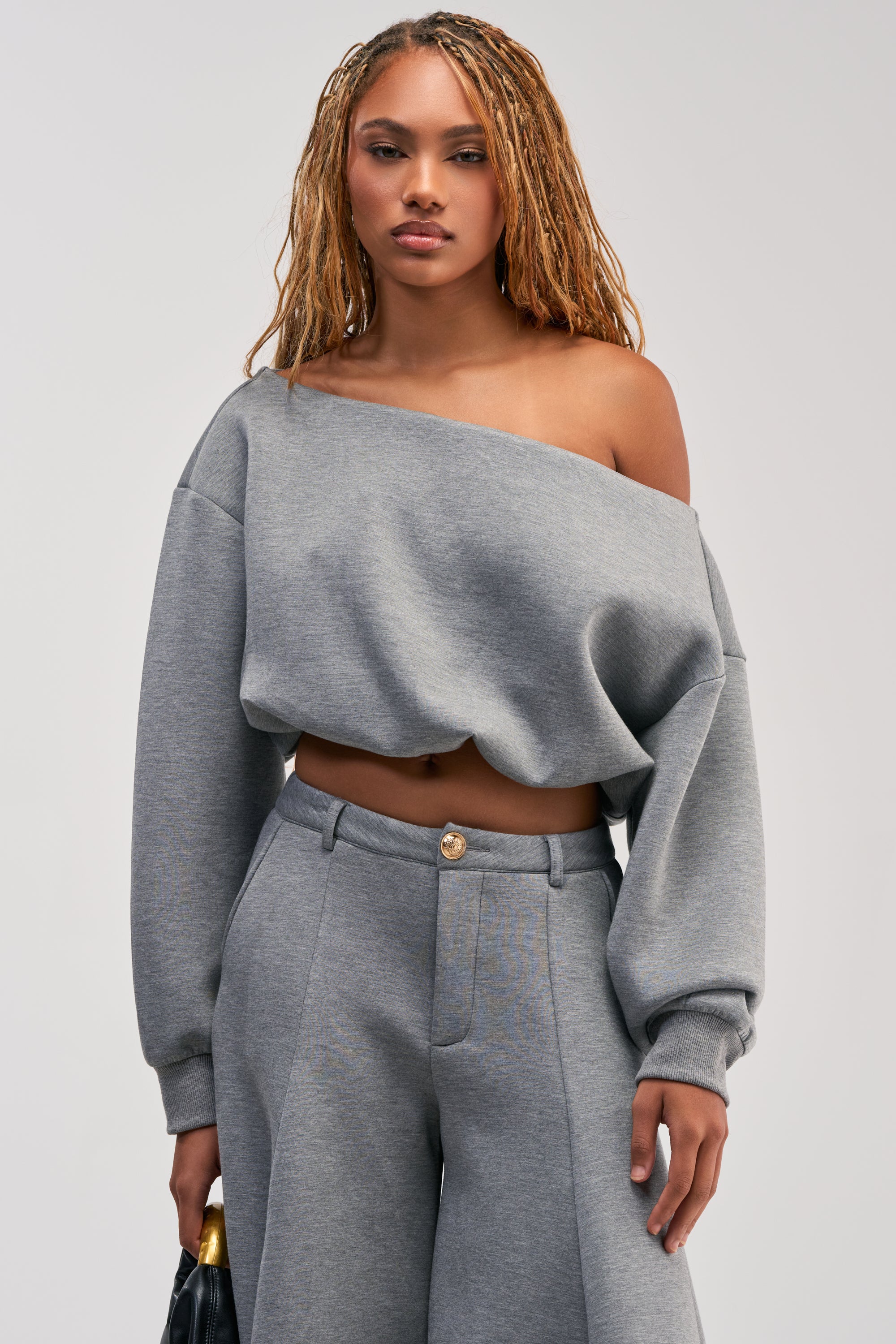 A woman with long blonde braids models the WILD AND FREE OFF THE SHOULDER TOP—a grey, cropped sweatshirt that’s a modern streetwear essential—styled with matching high-waisted pants and a black handbag, standing against a plain light background.