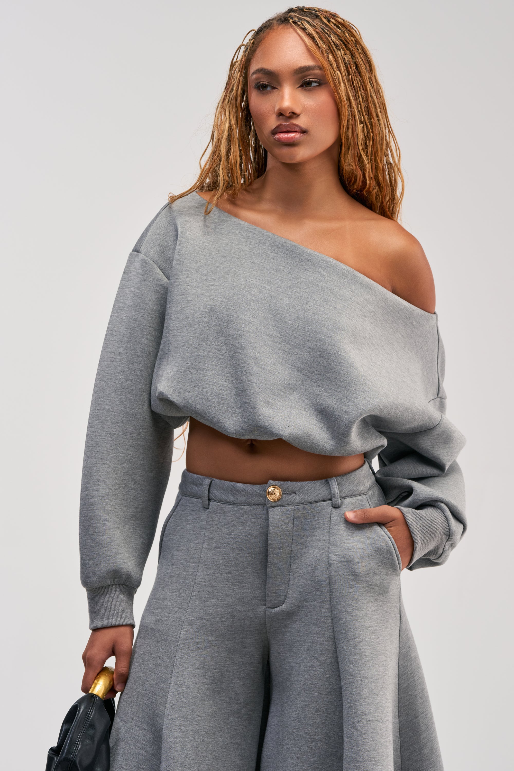 Wearing the WILD AND FREE OFF THE SHOULDER TOP in grey, paired with matching wide-leg pants, a woman stands confidently with one hand in her pocket and a black bag in the other, channeling modern streetwear style as she gazes to the side.