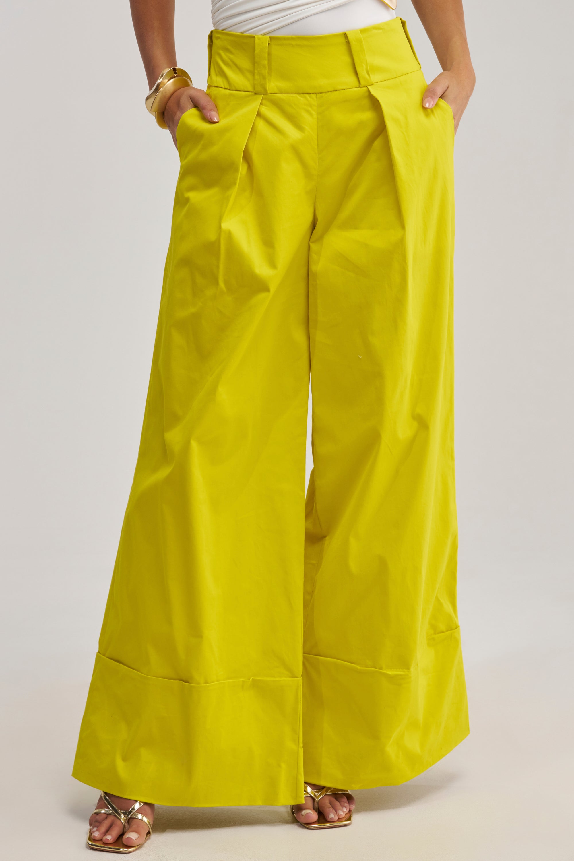 Wearing the GIVE IT TO ME BABY TROUSER in yellow with wide legs and pleats, gold sandals, a white top, and gold bracelets, this person stands hands-in-pockets against a plain background—effortlessly funky style.
