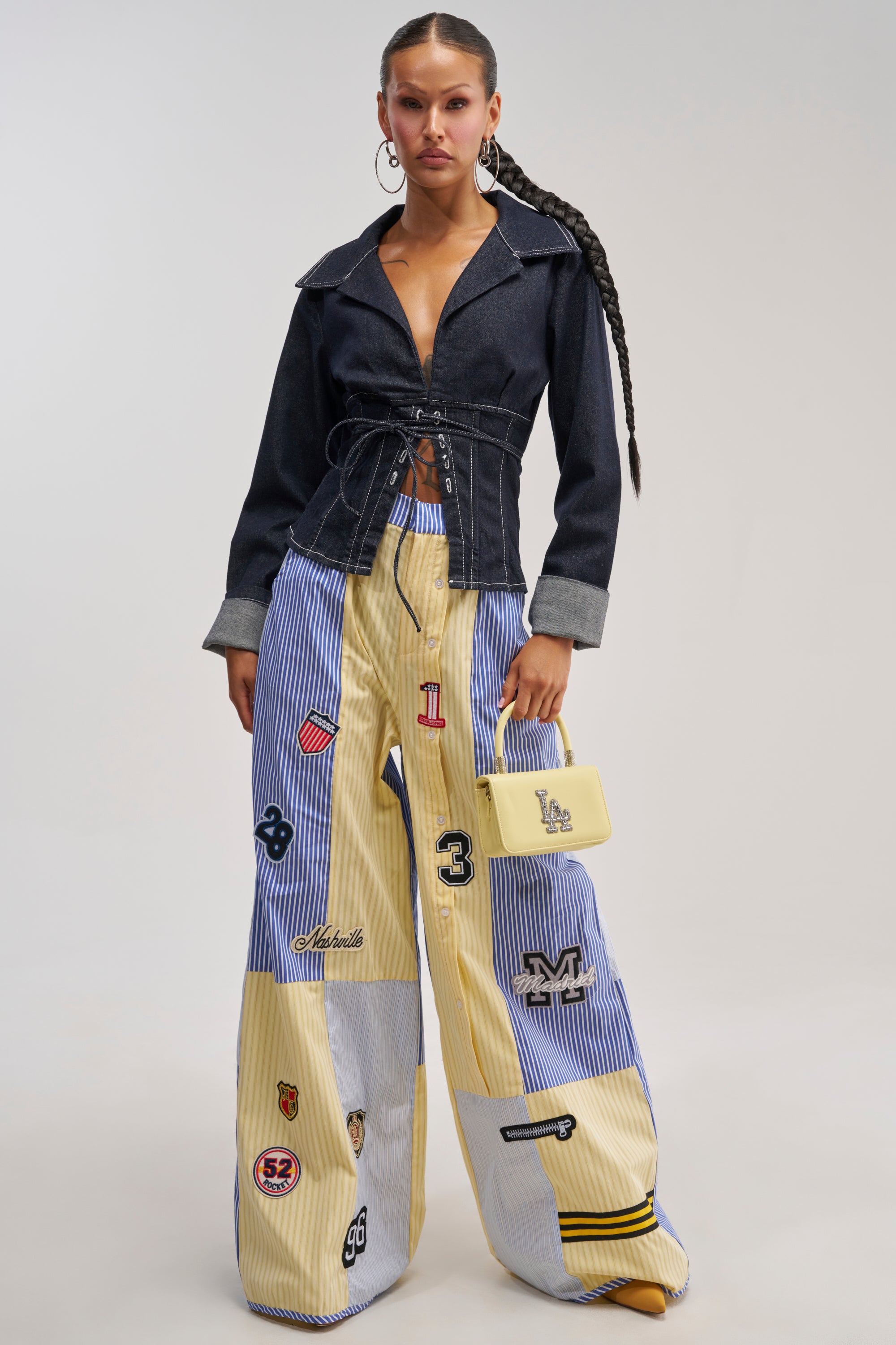 A woman wears wide-leg BACK TO SCHOOL POPLIN TROUSER featuring patchwork stripes and varsity-inspired details, paired with a dark lace-up top. She completes the look with a small yellow handbag and long braids, standing against a neutral backdrop to capture an effortlessly stylish streetwear vibe.
