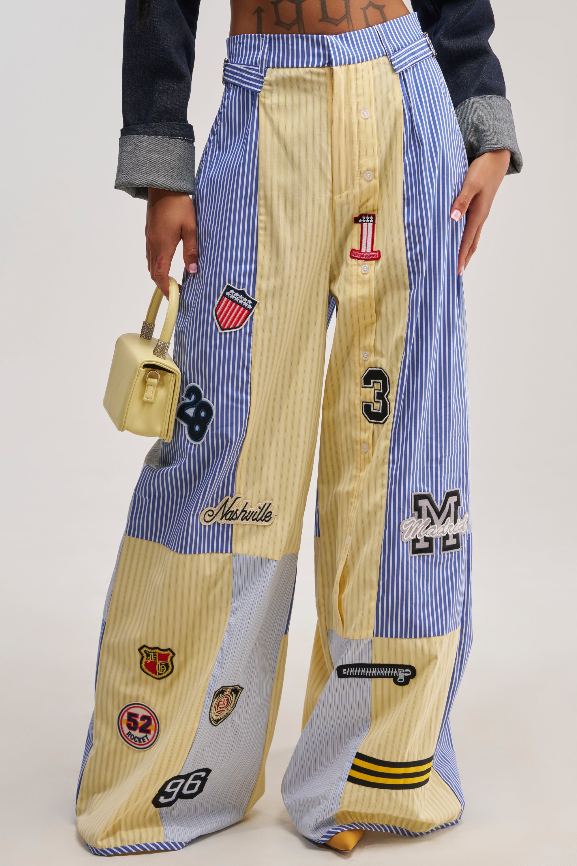 A person wearing the BACK TO SCHOOL POPLIN TROUSER, characterized by wide-leg patchwork stripes in blue and yellow and detailed with multiple varsity patches, paired with a small yellow handbag—a perfect showcase of bold streetwear styling. Only the lower torso and legs are visible.