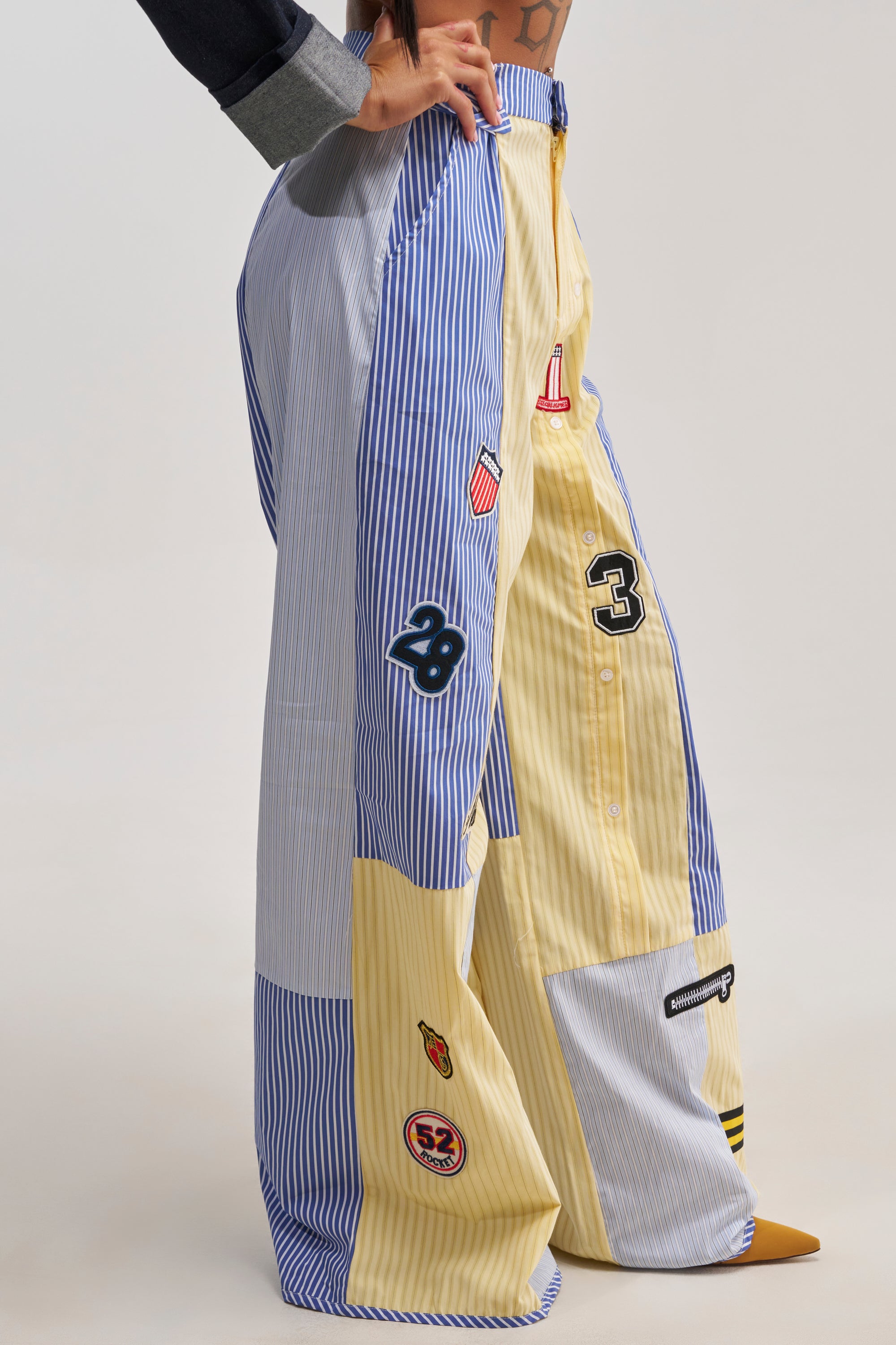A person is shown standing on a plain background wearing the BACK TO SCHOOL POPLIN TROUSER—wide-leg patchwork stripe poplin trousers featuring blue, white, and yellow stripes, adorned with varsity-inspired patches with numbers and small graphics. The upper body is not visible, evoking a modern streetwear vibe.