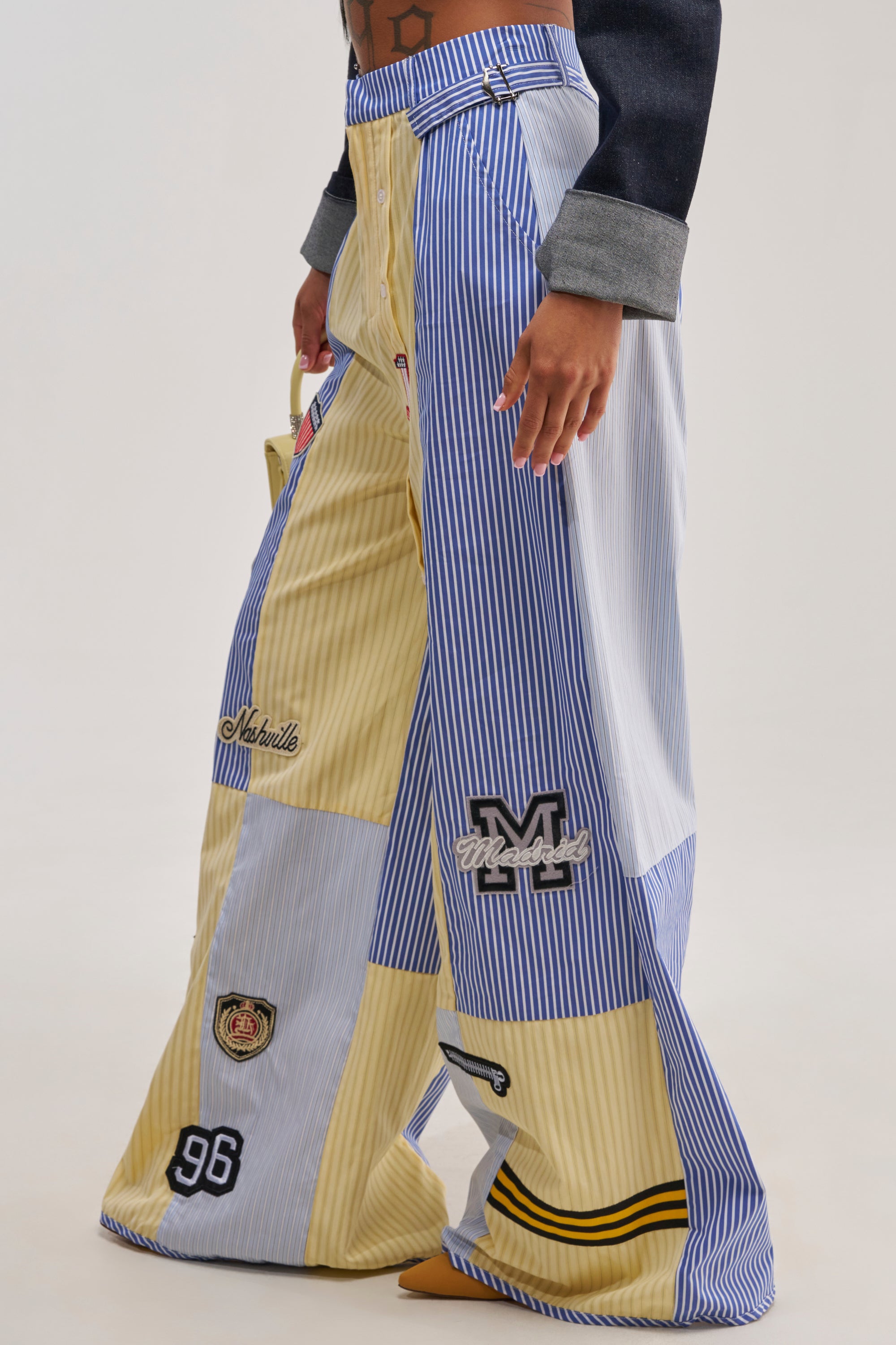 A person stands against a plain background wearing the BACK TO SCHOOL POPLIN TROUSER—wide-leg trousers crafted from various yellow, blue, and white striped poplin fabrics and featuring embroidered patches.