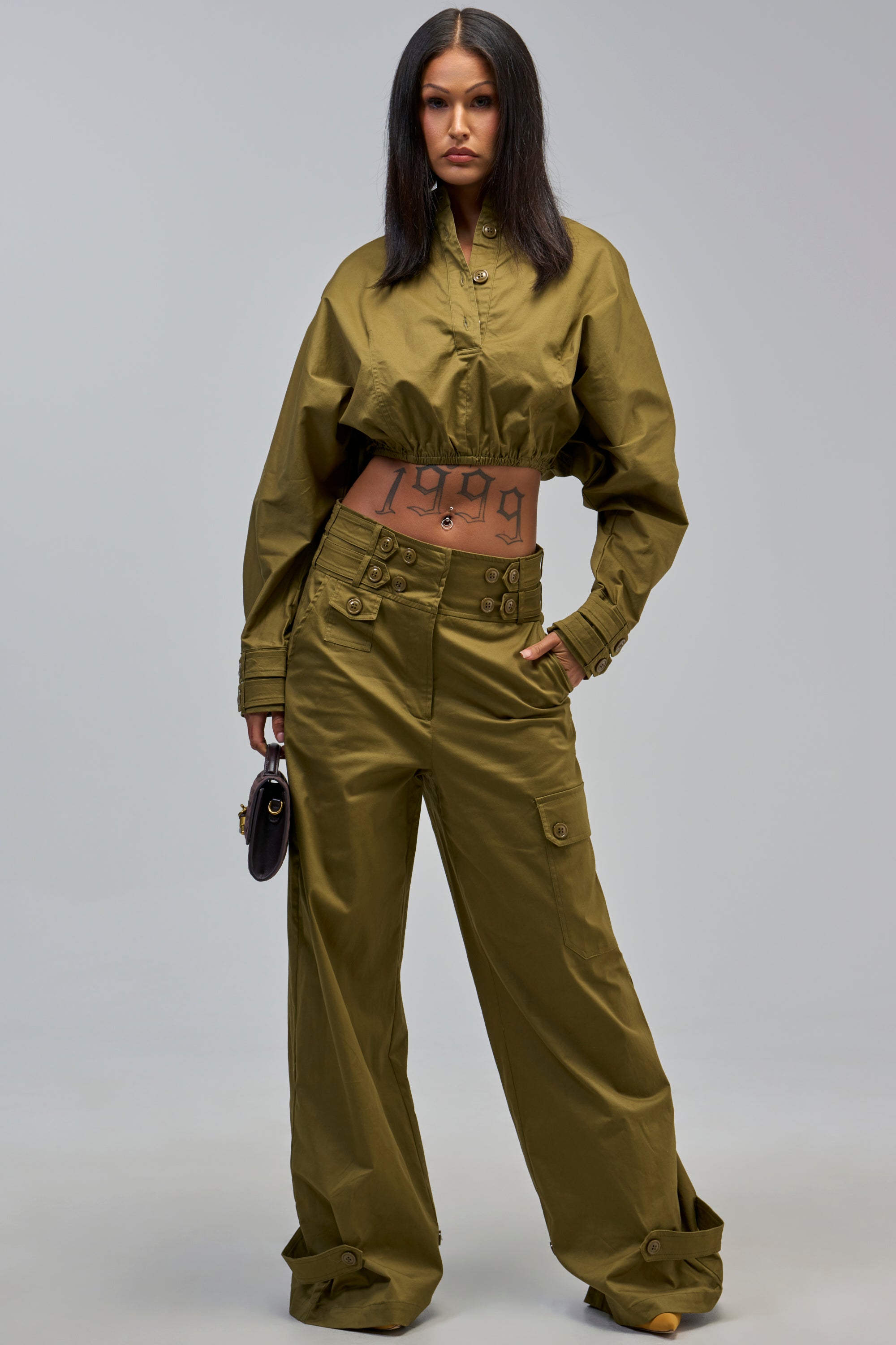 A woman stands confidently in olive green, wearing the HIGH FASHION TRENCH RELAXED PANT with cargo pockets and a cropped jacket. She holds a small black bag, showing her “1999” tattoo and long straight hair.