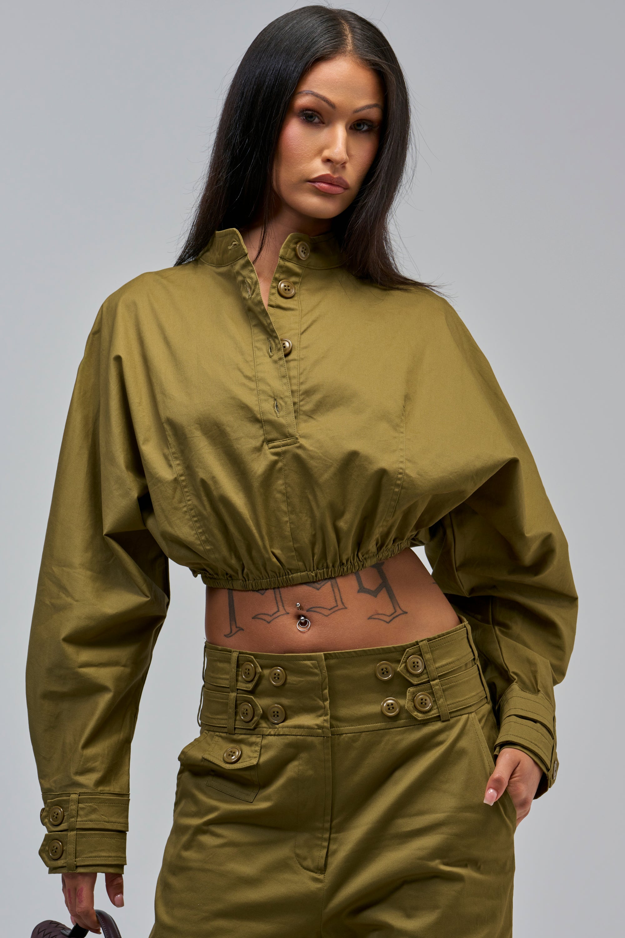 A woman with long dark hair stands confidently against a plain gray background, wearing the HIGH FASHION TRENCH TOP and matching pants that reveal geometric tattoos on her midriff.