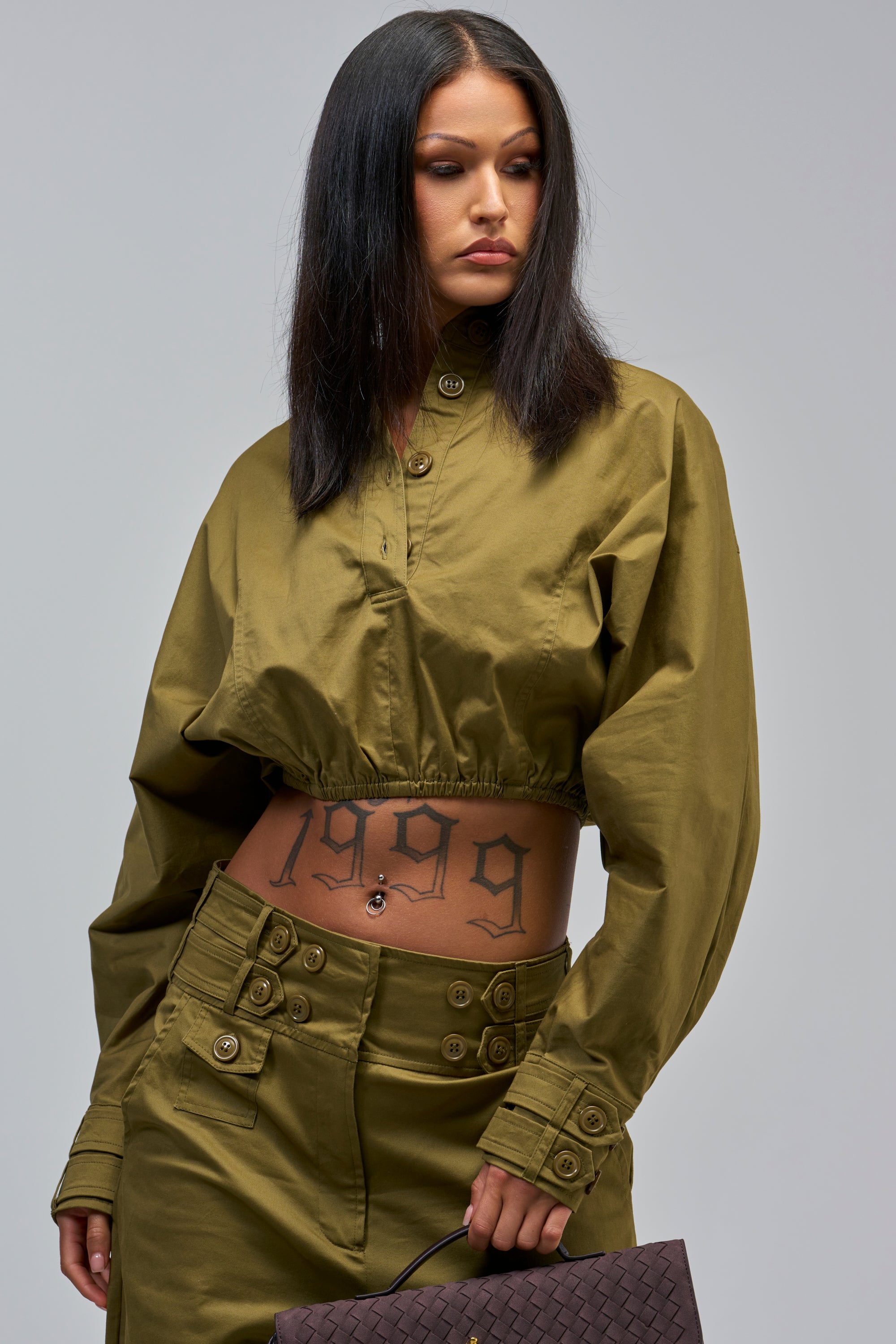 A woman with straight dark hair wears the HIGH FASHION TRENCH TOP in olive green with matching pants, revealing a “1999” tattoo on her stomach. She holds a textured dark handbag and looks to the side.