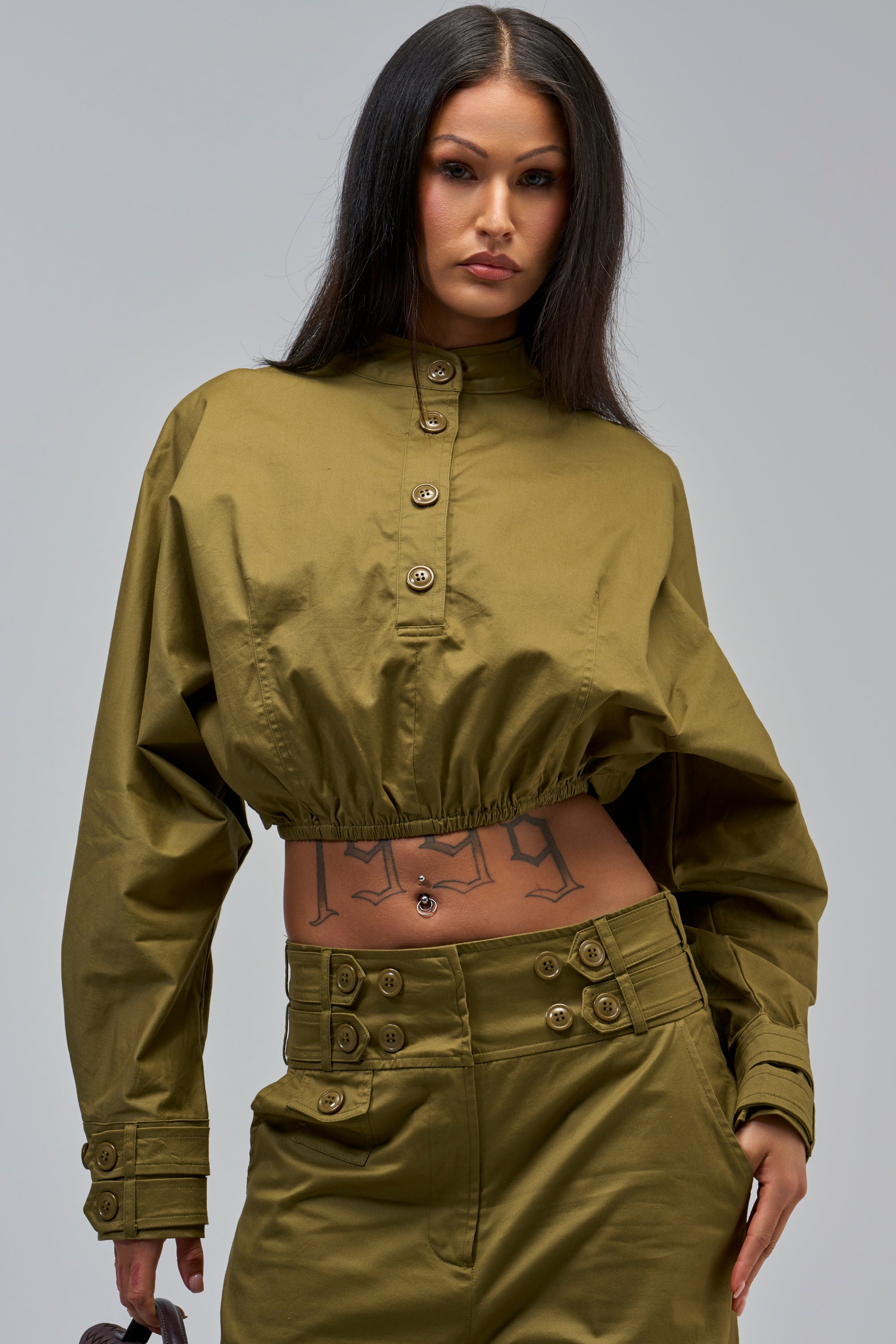A woman showcases the HIGH FASHION TRENCH TOP and matching high-waisted pants, revealing a stomach tattoo as she stands confidently against a plain gray background.
