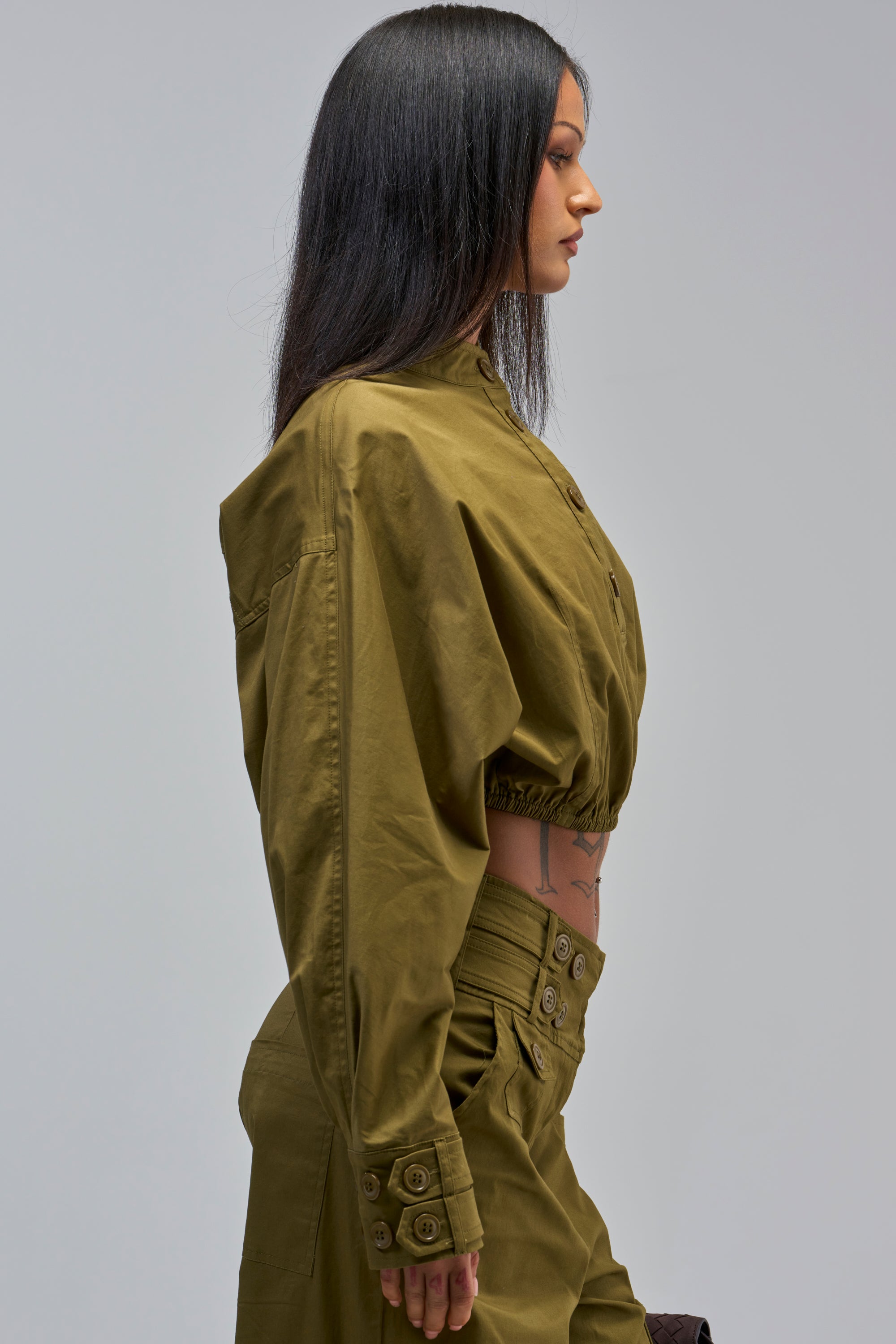 Woman in profile wears the HIGH FASHION TRENCH TOP in olive green with matching high-waisted pants. Her straight dark hair is down and a torso tattoo is visible, set against a plain light gray background.