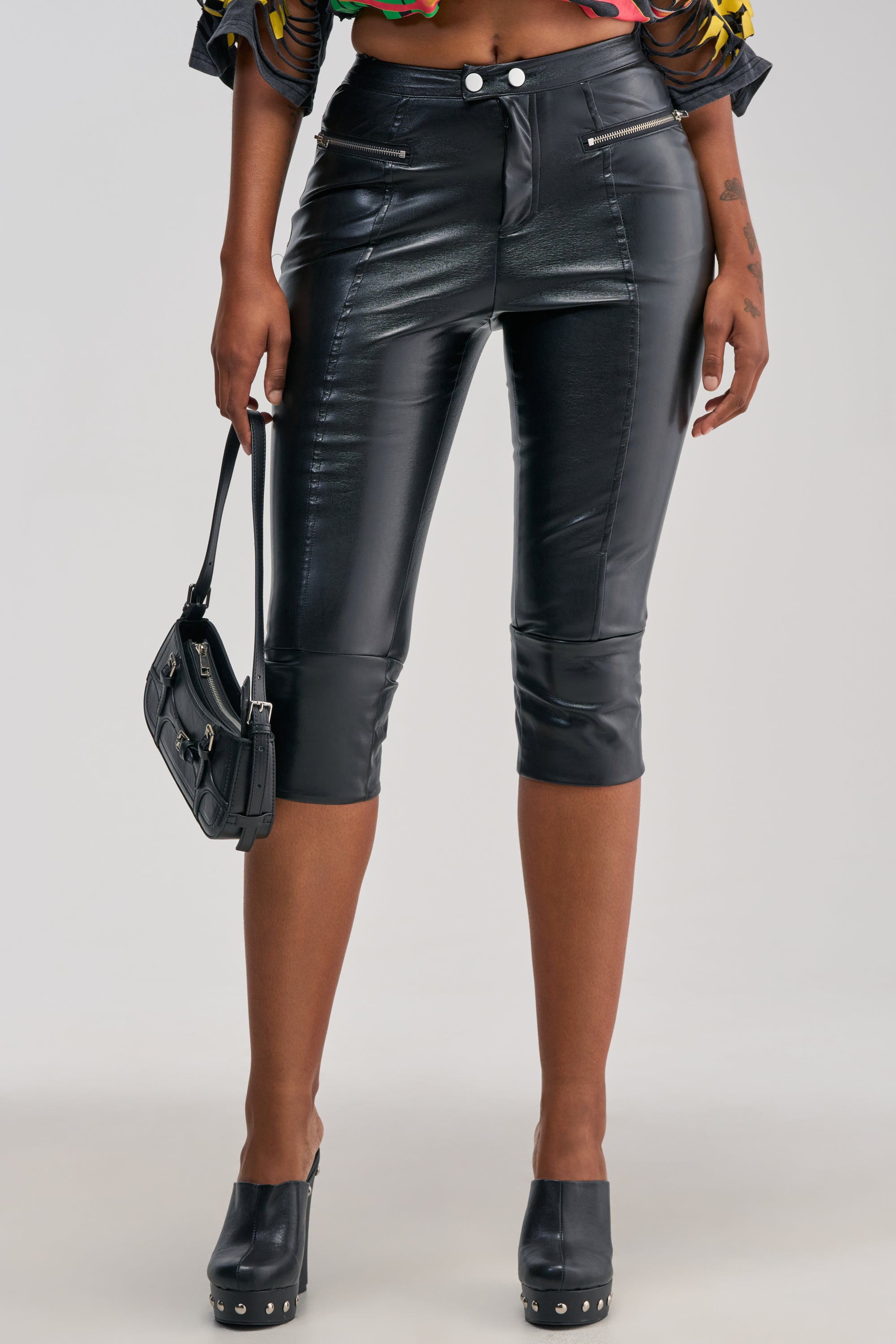 A person is shown from the shoulders down against a plain background, dressed in bold street style featuring the “A LITTLE BIT OF THIS FAUX LEATHER CAPRI” pants paired with black heeled sandals and holding a small black handbag. This striking look is perfect for concert outfits or making a statement on the streets.