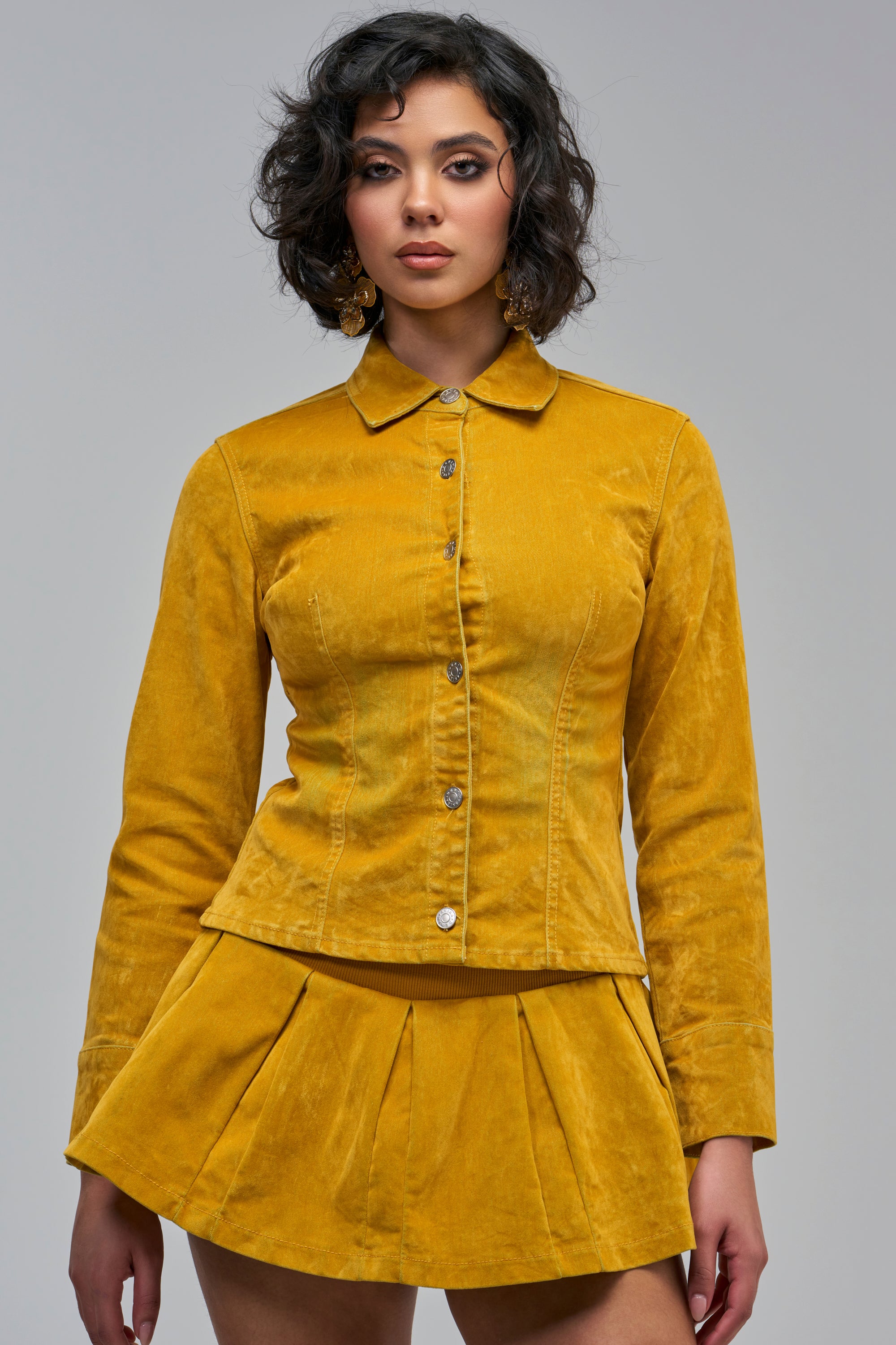 A person wearing a mustard yellow GOLDEN GIRL VELVET DENIM BUTTON DOWN jacket and a matching pleated mini skirt, perfect for fall fashion.
