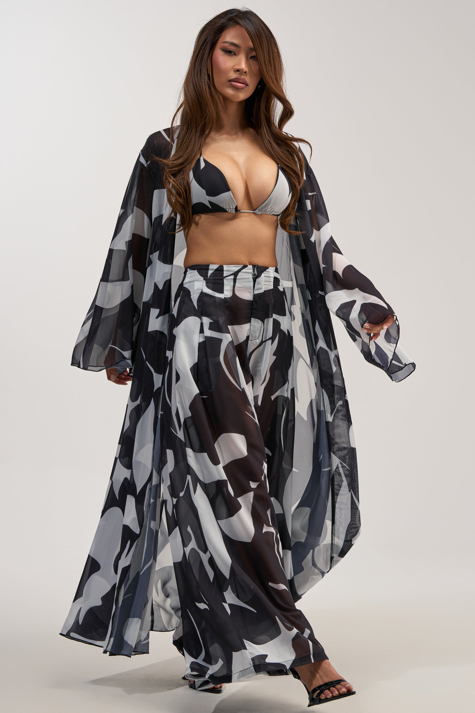 A woman models the BAECATION CHIFFON DUSTER—a three-piece set featuring a black, white, and gray abstract print bikini top, wide-leg pants, and a coordinating long sheer chiffon duster—styled with black high-heeled sandals against a plain backdrop for effortless resort wear.