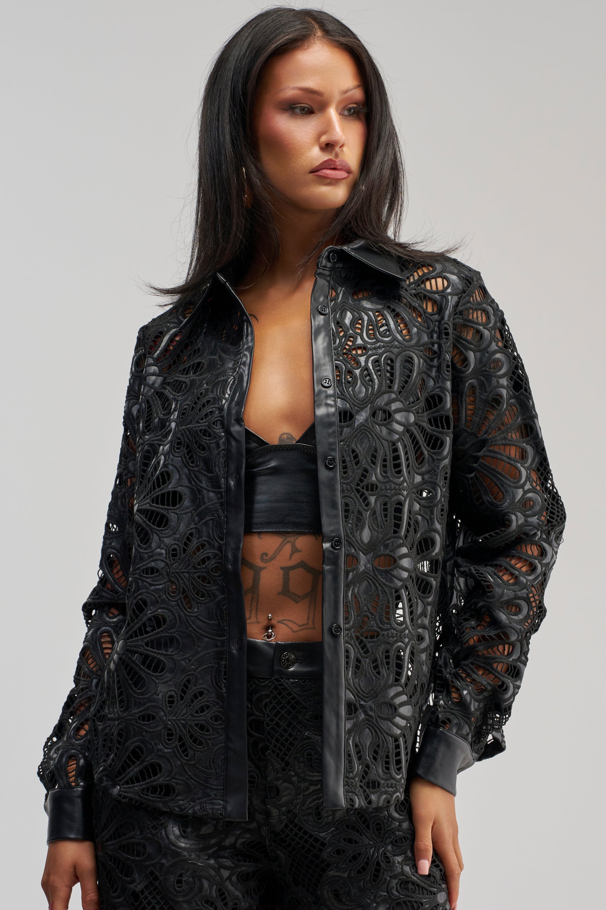 A woman is wearing the LUCKY ME LONG SLEEVE BUTTON DOWN—a black, patterned leather jacket with faux leather trim—styled over a black bandeau top and matching pants, her tattoos visible on her stomach as she stands against a neutral background.