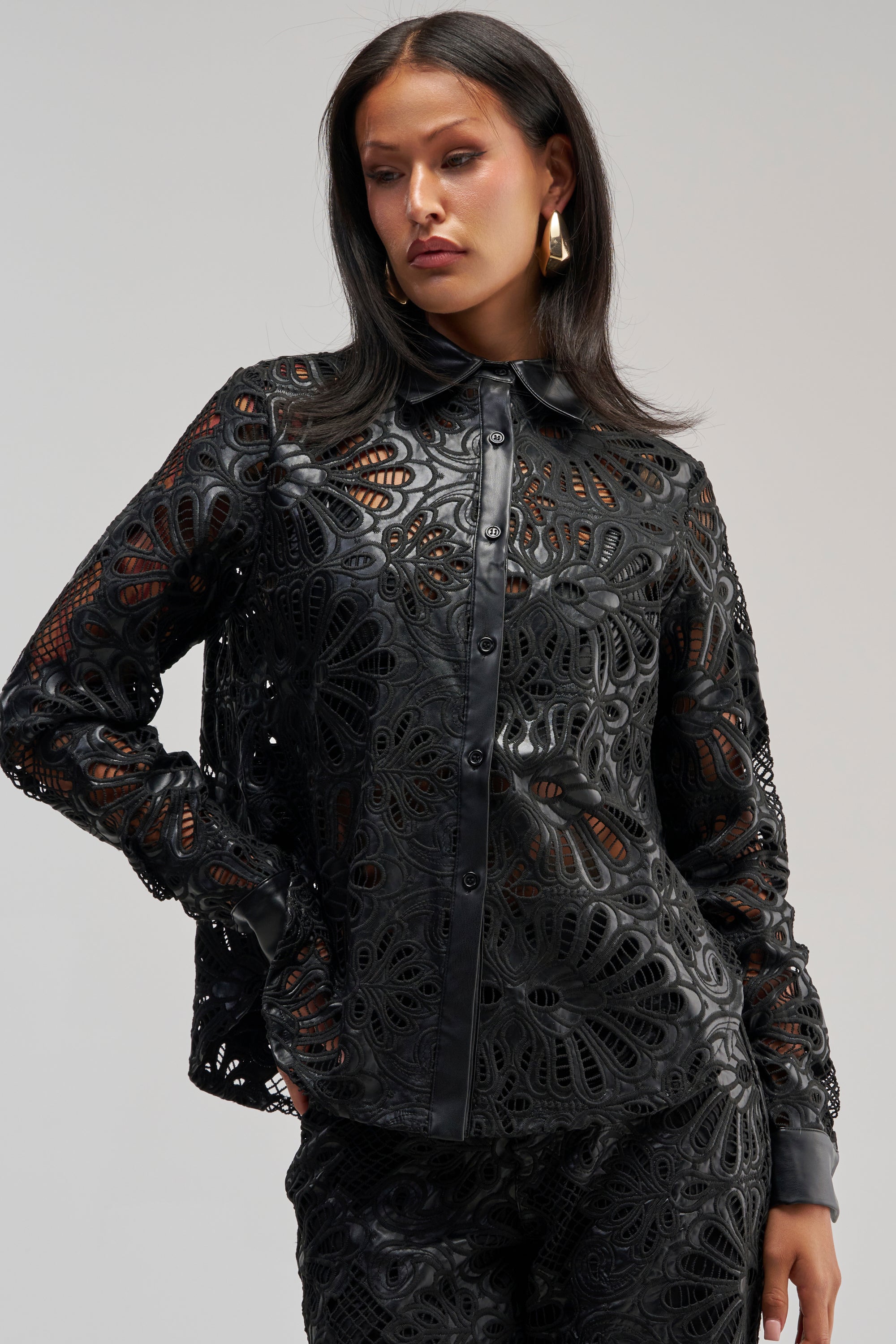 A woman poses in the LUCKY ME LONG SLEEVE BUTTON DOWN, a black blouse with faux leather trim, paired with matching pants featuring an intricate cutout pattern. She has long dark hair and wears large gold hoop earrings.