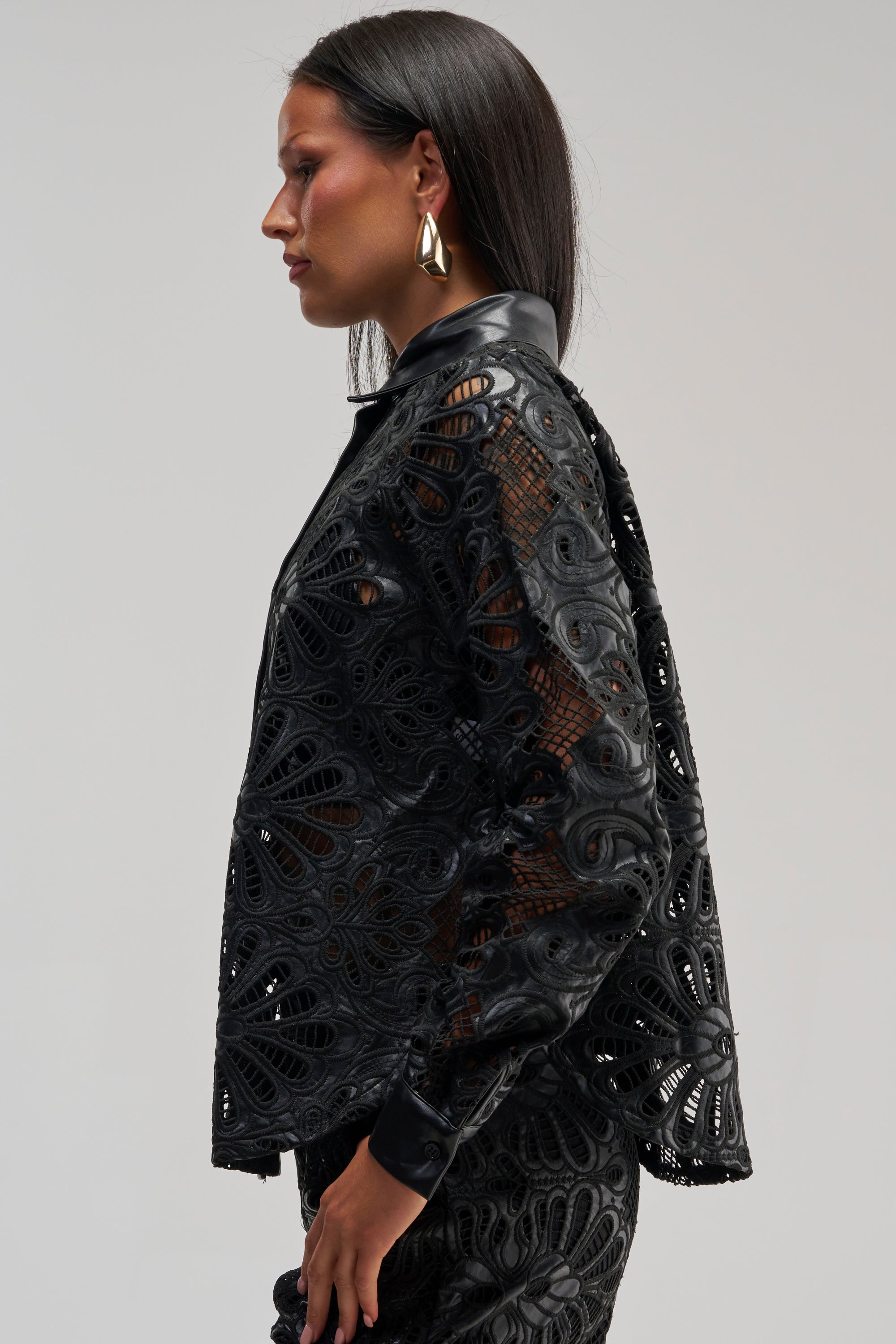 Facing left against a plain light background, a person with long dark hair wears the LUCKY ME LONG SLEEVE BUTTON DOWN—a black floral blouse featuring intricately patterned lace and faux leather trim—paired with gold geometric earrings.