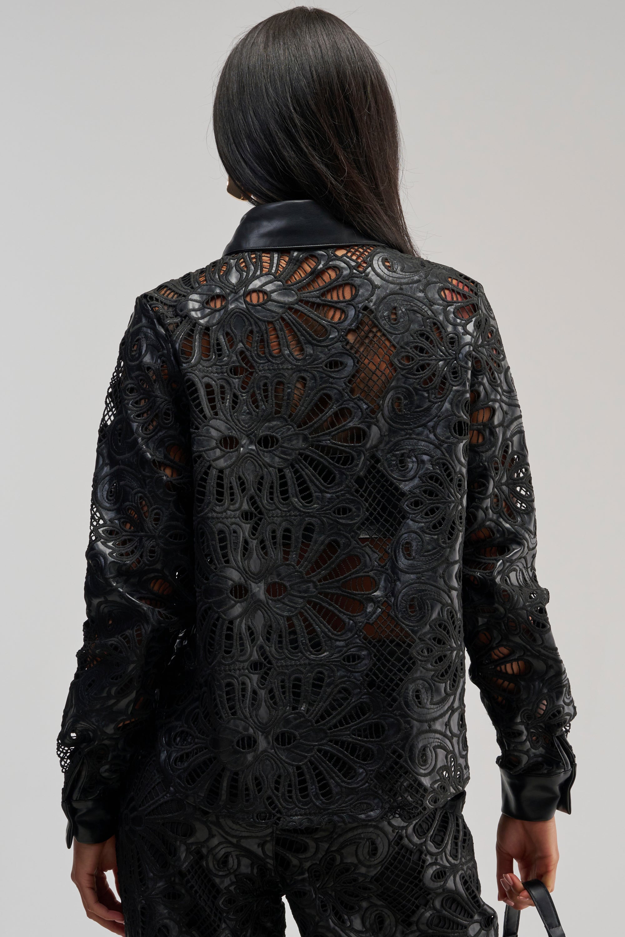 A person with long dark hair faces away, wearing a black, lace-patterned jacket with large floral designs and sheer sections layered over the LUCKY ME LONG SLEEVE BUTTON DOWN—a black floral blouse with faux leather trim—while holding a small black bag.