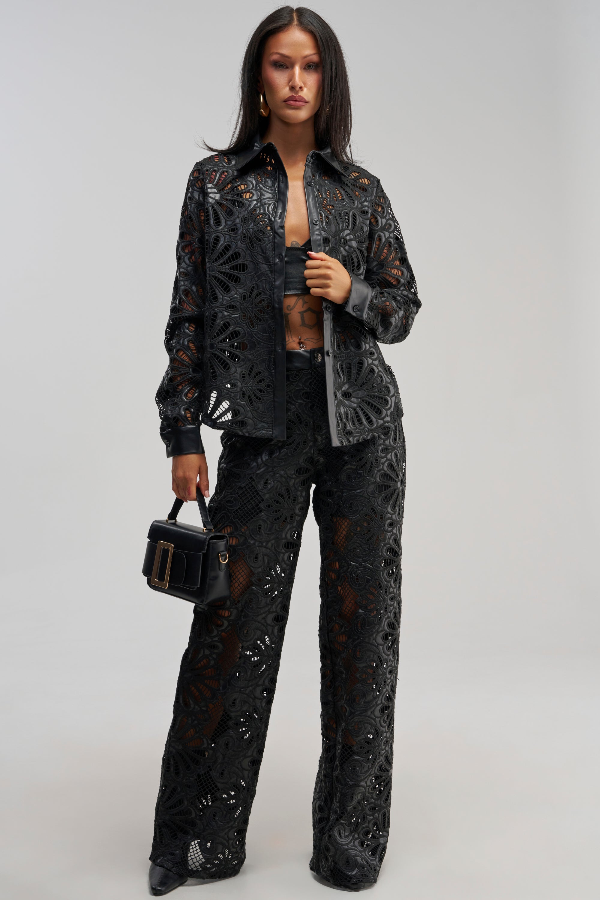 A woman stands against a plain, light-colored background wearing the LUCKY ME TROUSER—black lace wide-leg streetwear pants paired with a matching coordinating bralette and small black handbag. She has long straight hair and black pointed shoes.