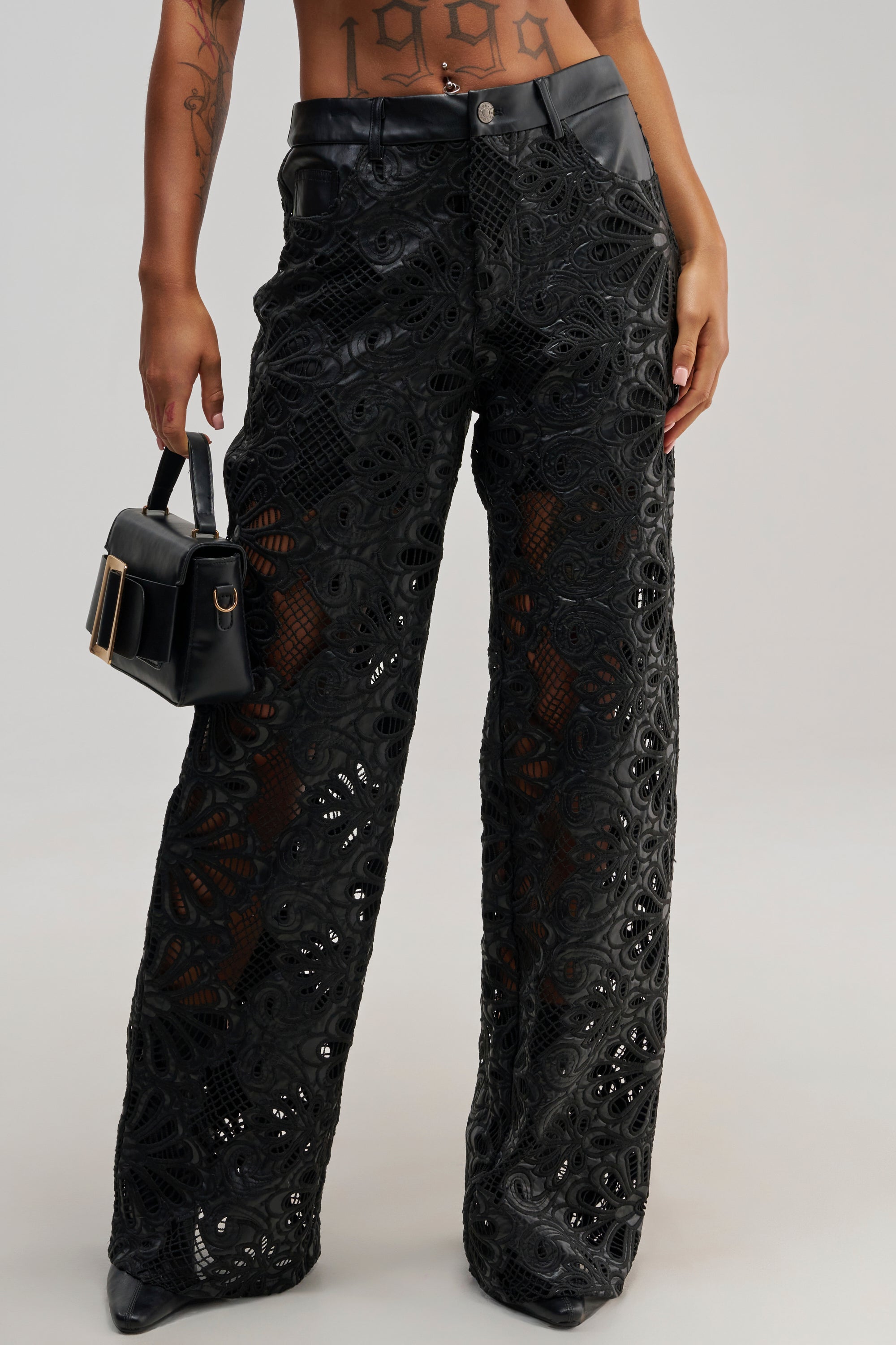 A person wearing the LUCKY ME TROUSER, featuring a high-waisted black lace wide-leg streetwear design with cutout details, and holding a small black handbag. Only the lower torso and legs are visible.