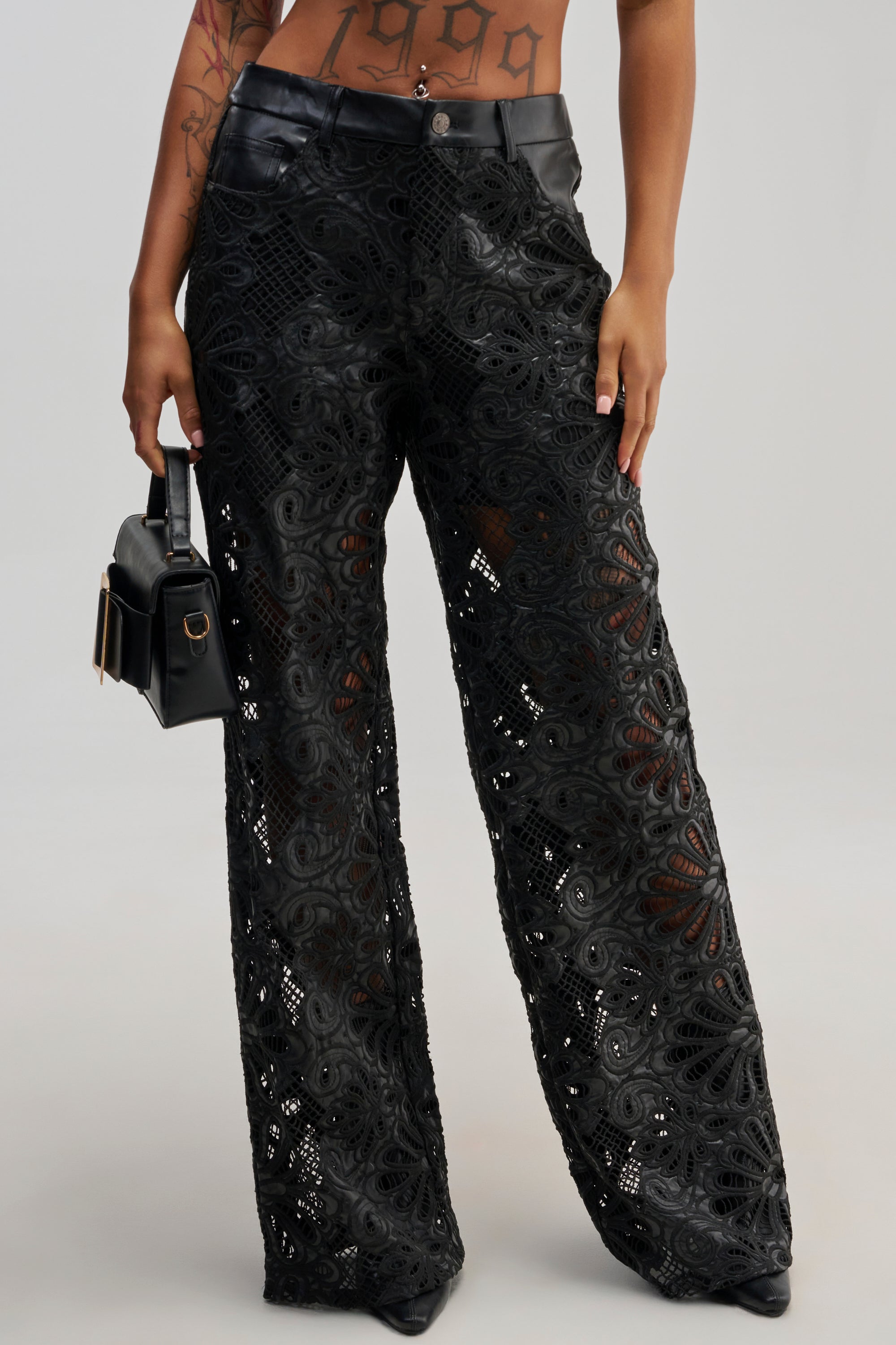 Wearing the LUCKY ME TROUSER—a pair of black lace floral overlay trousers with a wide-leg cut and semi-sheer finish, accented by a sleek faux leather waistband—the person holds a black handbag. Their midriff is visible, revealing a “1999” tattoo.
