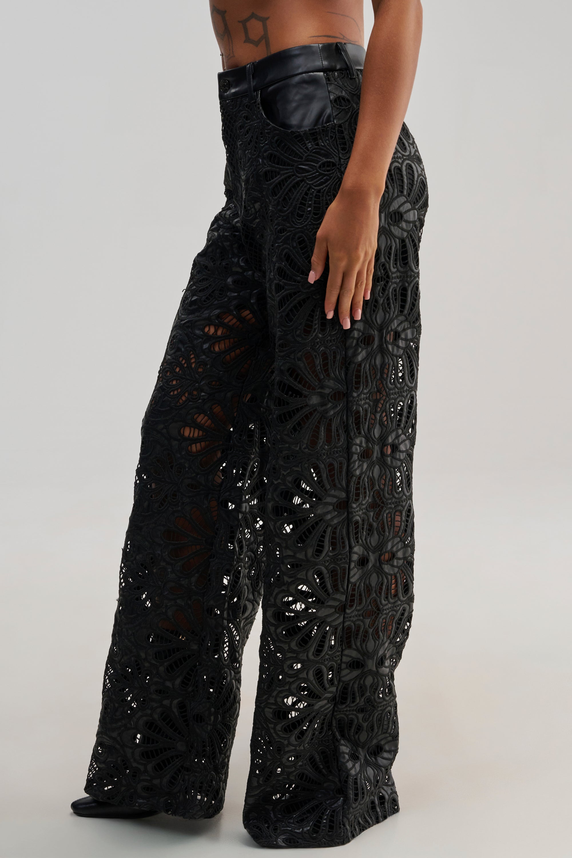 A model wearing the LUCKY ME TROUSER, a high-waisted wide-leg streetwear pant in black lace with a floral cutout pattern and leather details, stands against a plain light background.