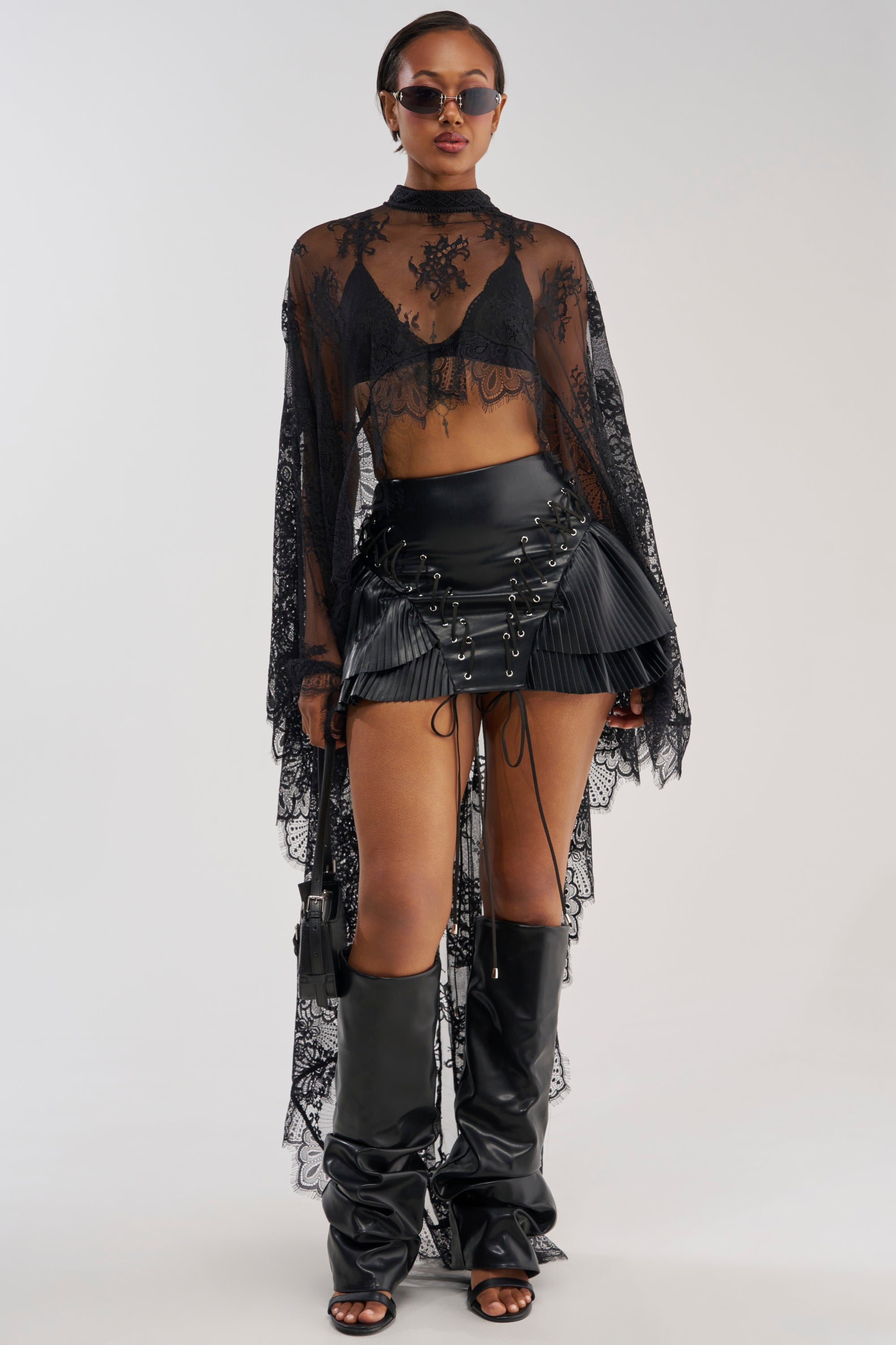 A woman stands against a plain background wearing sunglasses, a sheer black lace top over a black bralette, tall slouchy black boots, and carrying a small black purse. She completes her outfit with the COME FIND ME FAUX LEATHER MINI SKIRT featuring lace-up details.