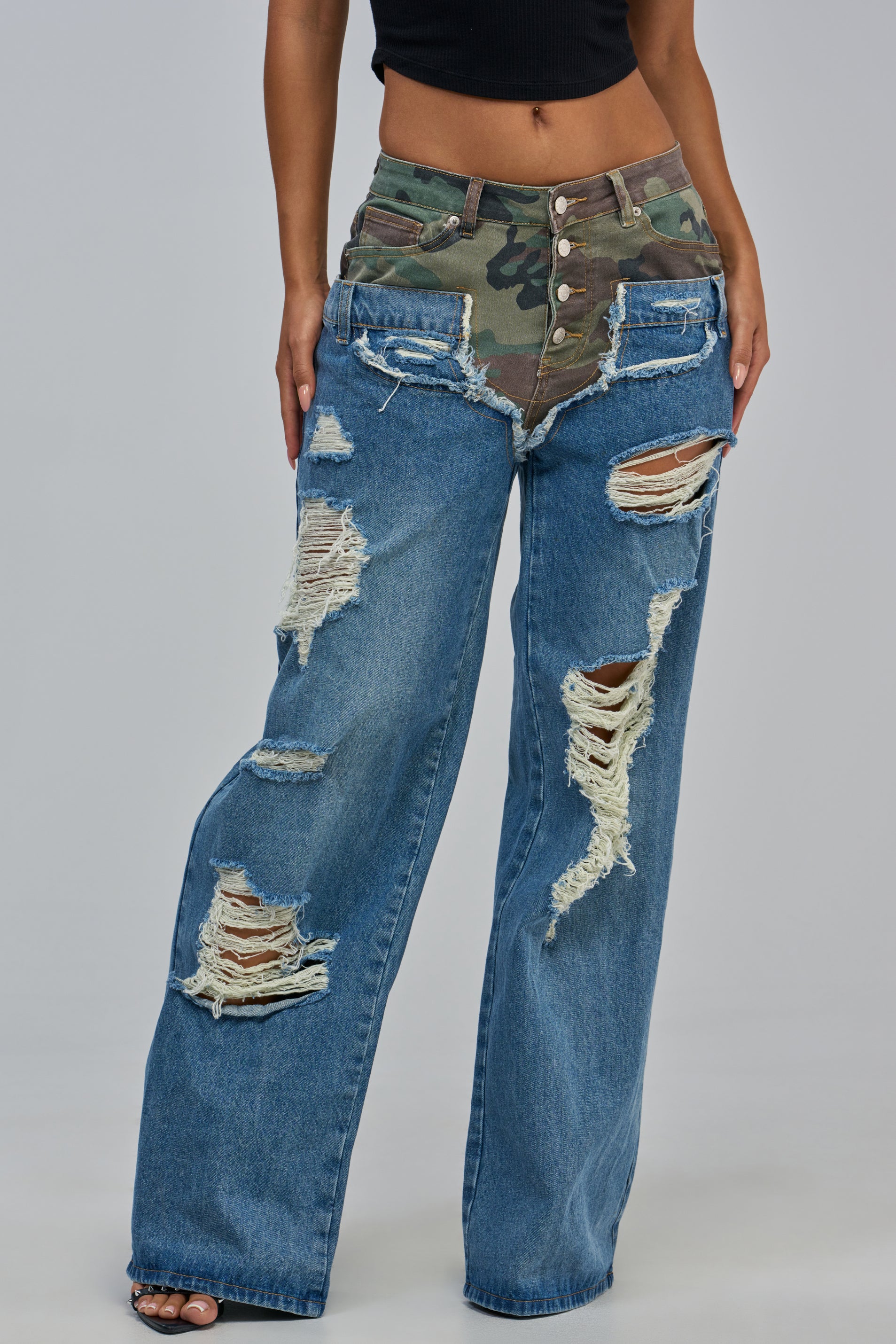 A person is shown wearing the “ITS GIVING RELAXED DENIM” jeans—medium blue, wide-leg silhouette, featuring a camo-print waist and large distressed holes—paired with a black crop top. Only the lower torso and legs are visible.