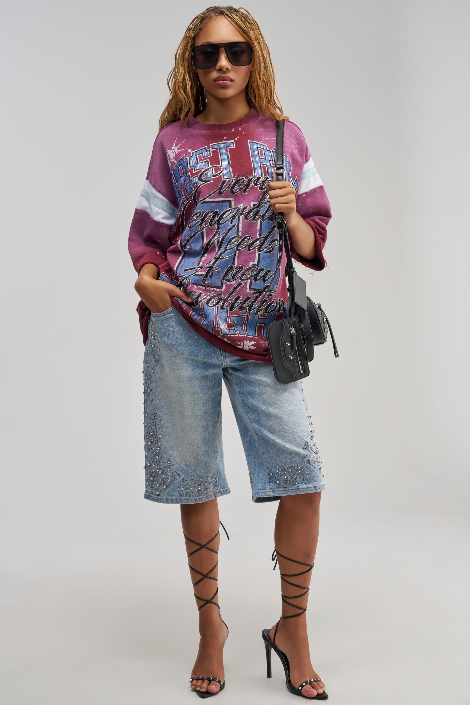 Wearing the BLING CITY BERMUDA SHORT, a woman in sunglasses pairs sparkling rhinestone Bermuda shorts with a loose maroon graphic shirt, strappy black high heels, and a black purse. She stands against a plain light background with her hand in her pocket.