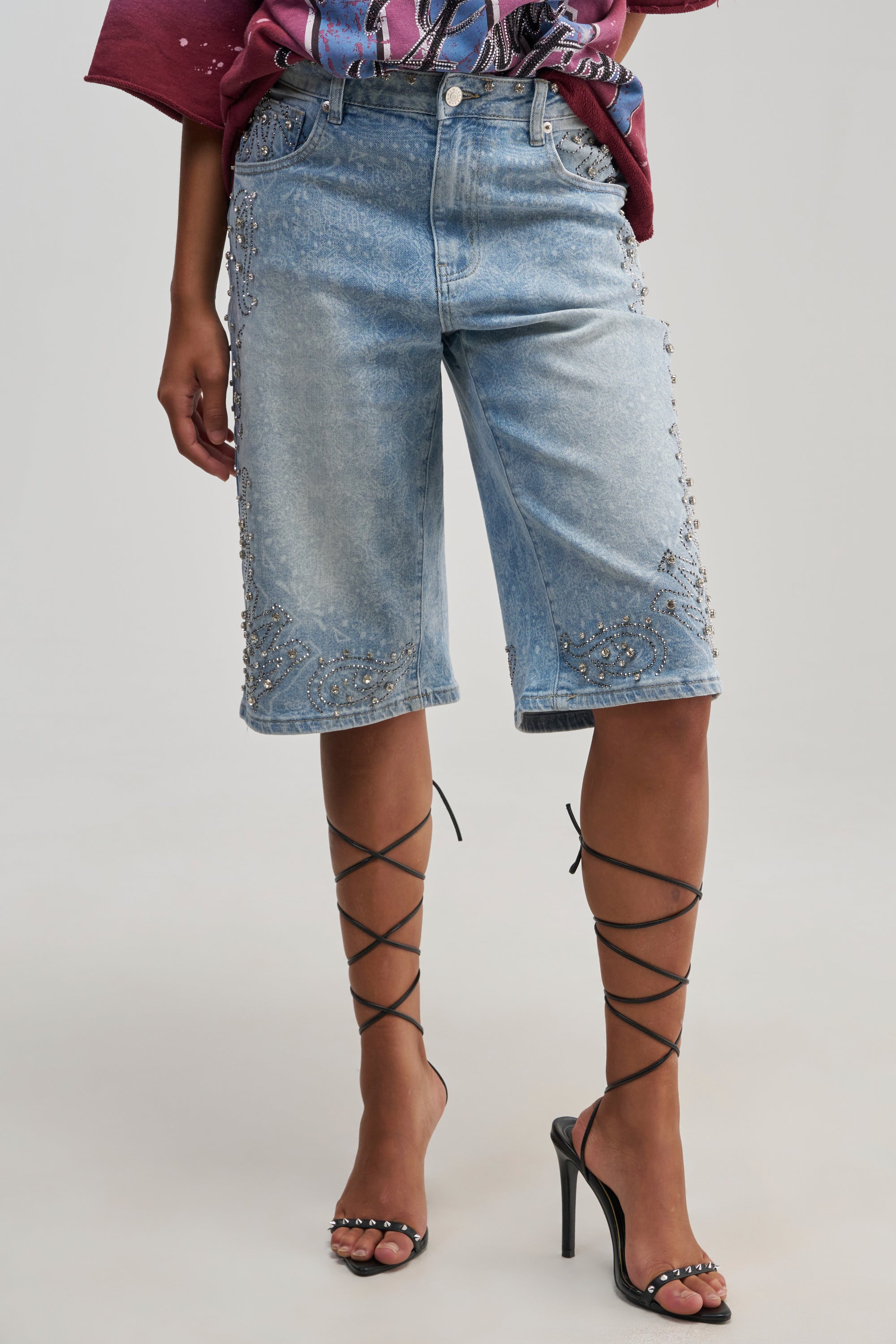 A person is wearing the BLING CITY BERMUDA SHORT—knee-length, light blue denim shorts embellished with rhinestones and silver studs—paired with a maroon shirt and black high-heeled sandals featuring crisscross straps up the calves. The background is plain and light-colored.